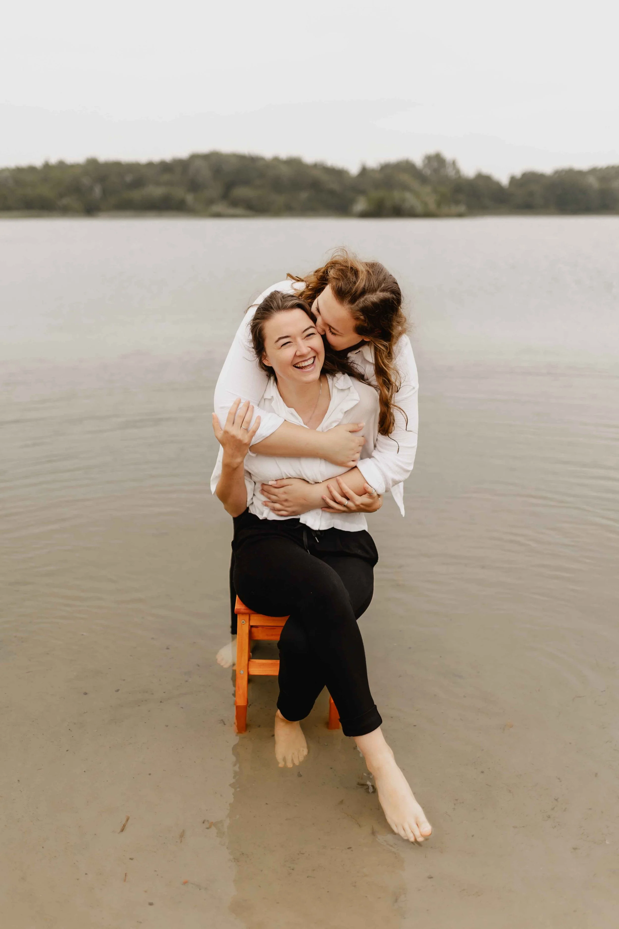 Zwei Frauen sitzen eng umschlungen auf einem kleinen Hocker im seichten Wasser eines Sees, lachend und voller Zuneigung
