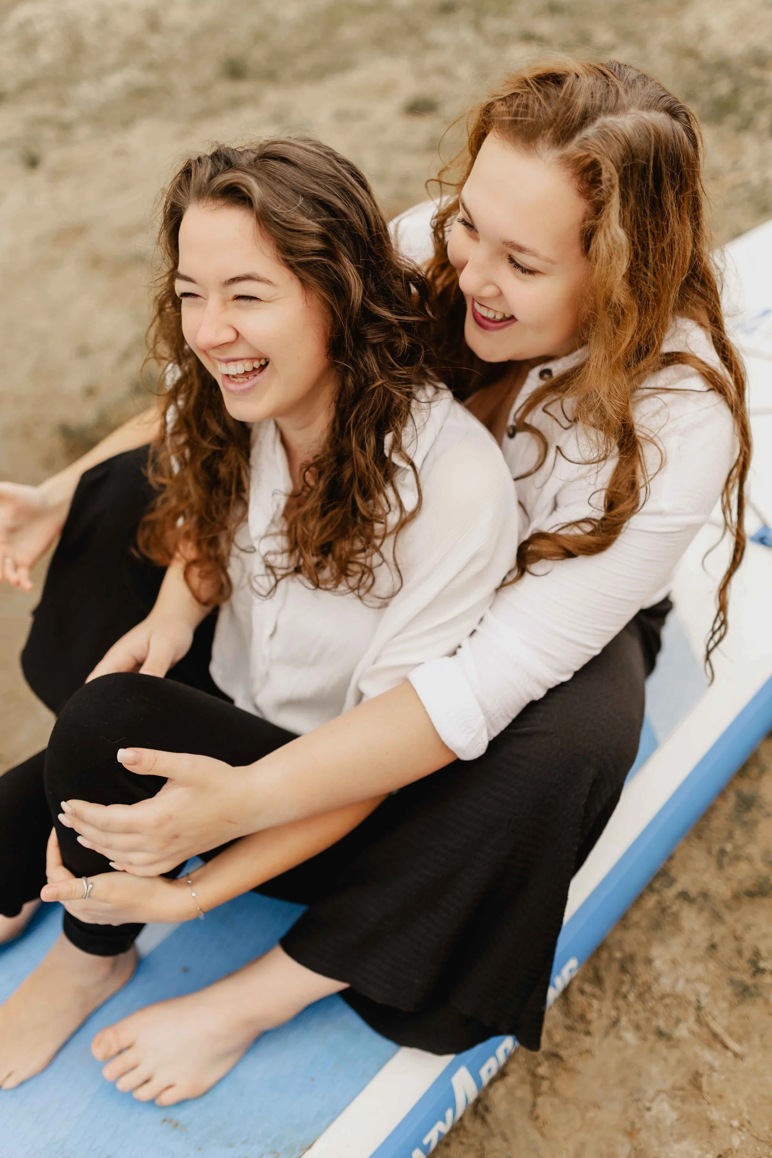 Zwei Frauen sitzen lachend auf einem Surfbrett am Oldenstädter See und genießen den Moment
