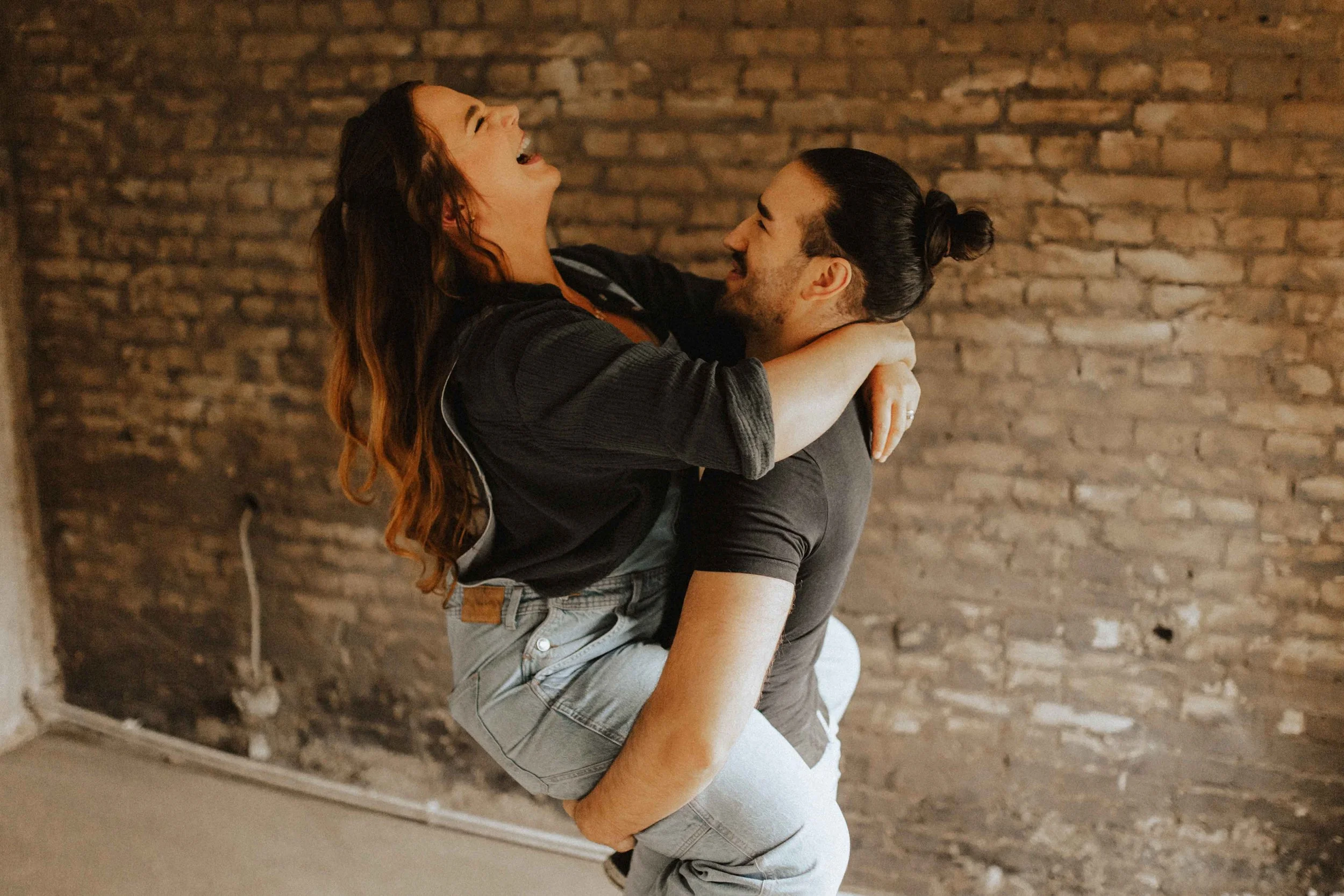 Paarshooting auf Baustelle – lachende Frau wird vom Mann hochgehoben und innig umarmt vor Backsteinwand in Lüneburg.