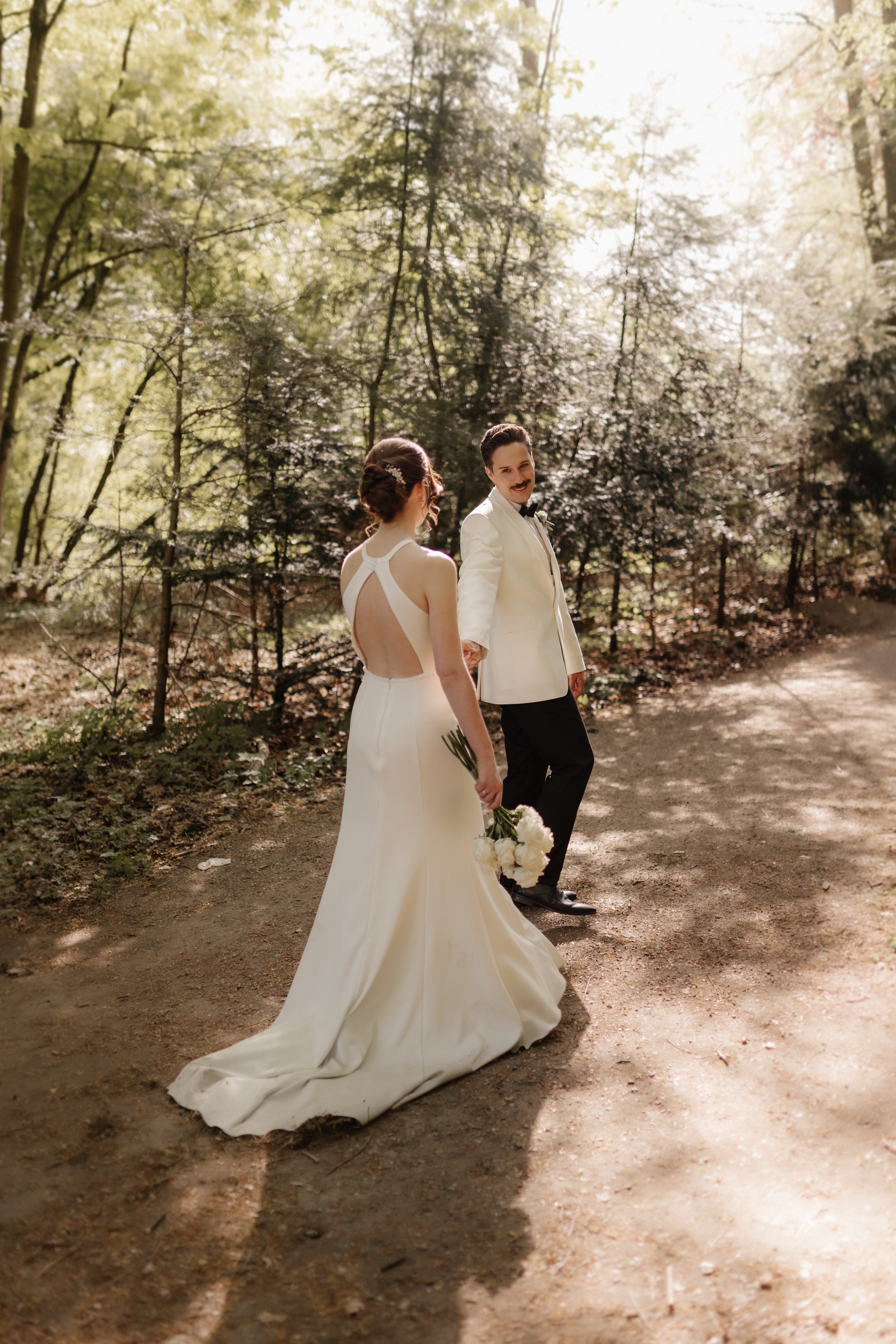 Brautpaar spaziert Hand in Hand über einen sonnigen Waldweg bei Lüneburg. Die Braut trägt ein elegantes Hochzeitskleid mit langer Schleppe und hält einen weißen Blumenstrauß.
