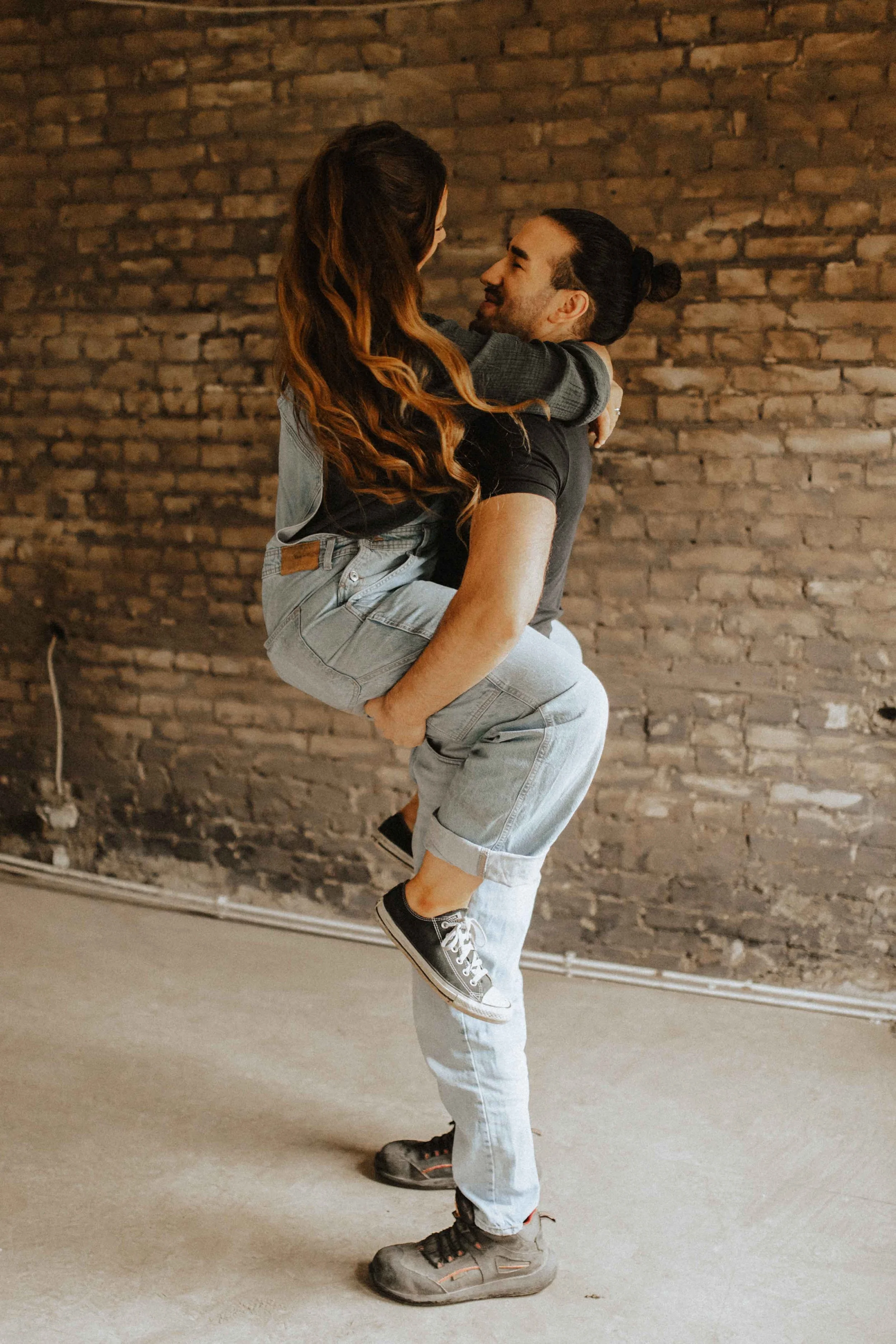 Verspieltes Paarshooting in Lüneburg – Mann hebt seine Partnerin hoch, beide blicken sich lächelnd in die Augen vor einer Backsteinwand.