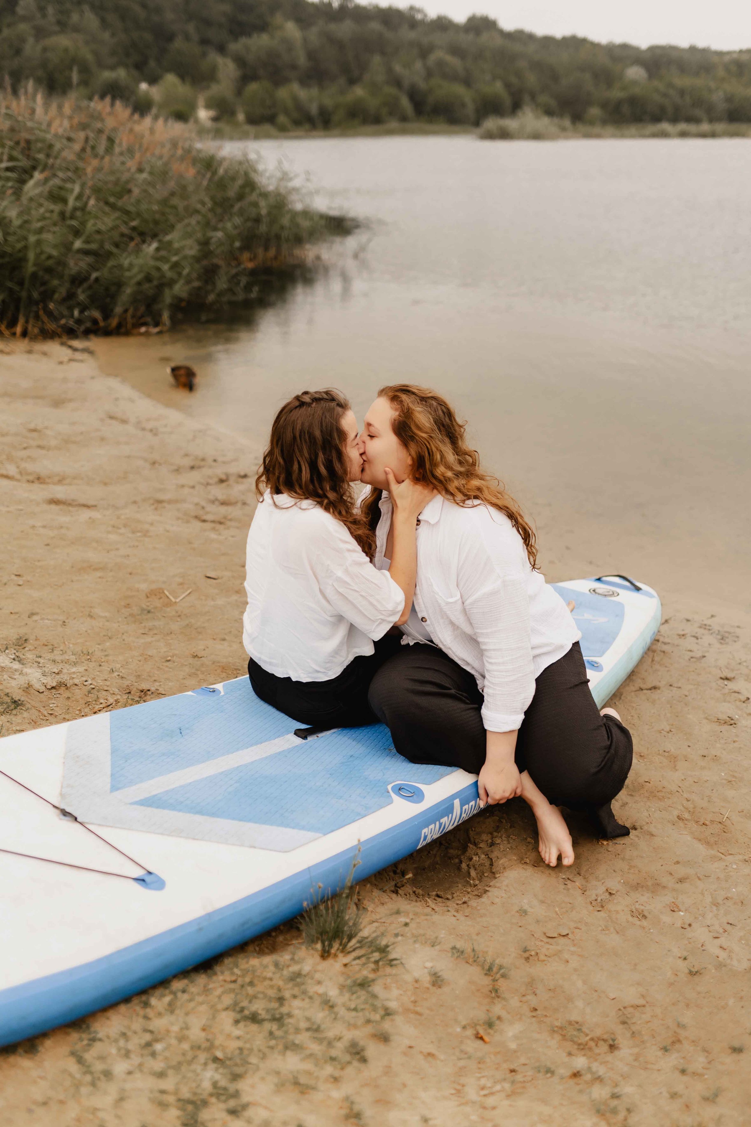 Zwei Frauen sitzen auf einem SUP-Board am Ufer des Oldenstädter Sees und küssen sich