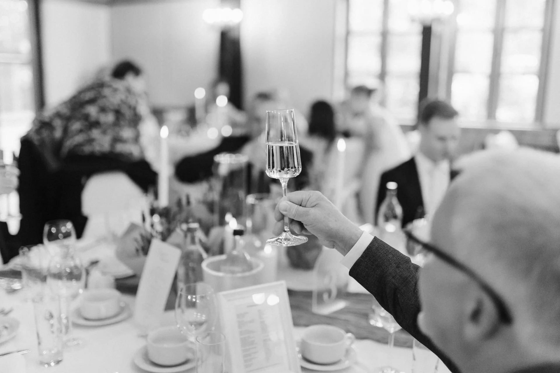 Schwarz-Weiß-Aufnahme eines Hochzeitsgastes, der mit einem Sektglas beim festlichen Hochzeitsempfang im Heide Hotel Reinstorf bei Lüneburg anstößt.