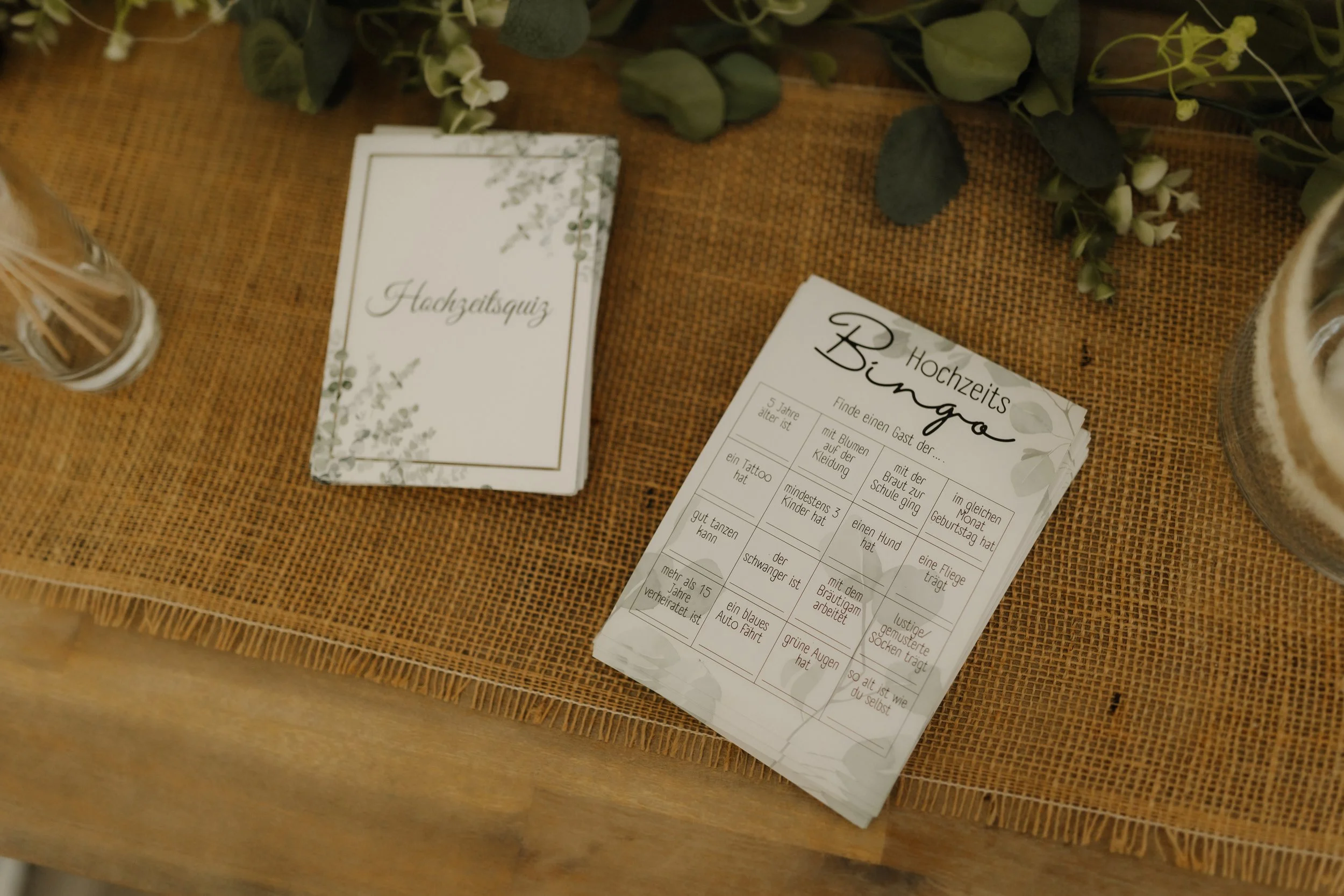 Hochzeitsbingo und liebevoll gestaltete Menükarten auf einem rustikalen Jutetischläufer mit grüner Blumendekoration.