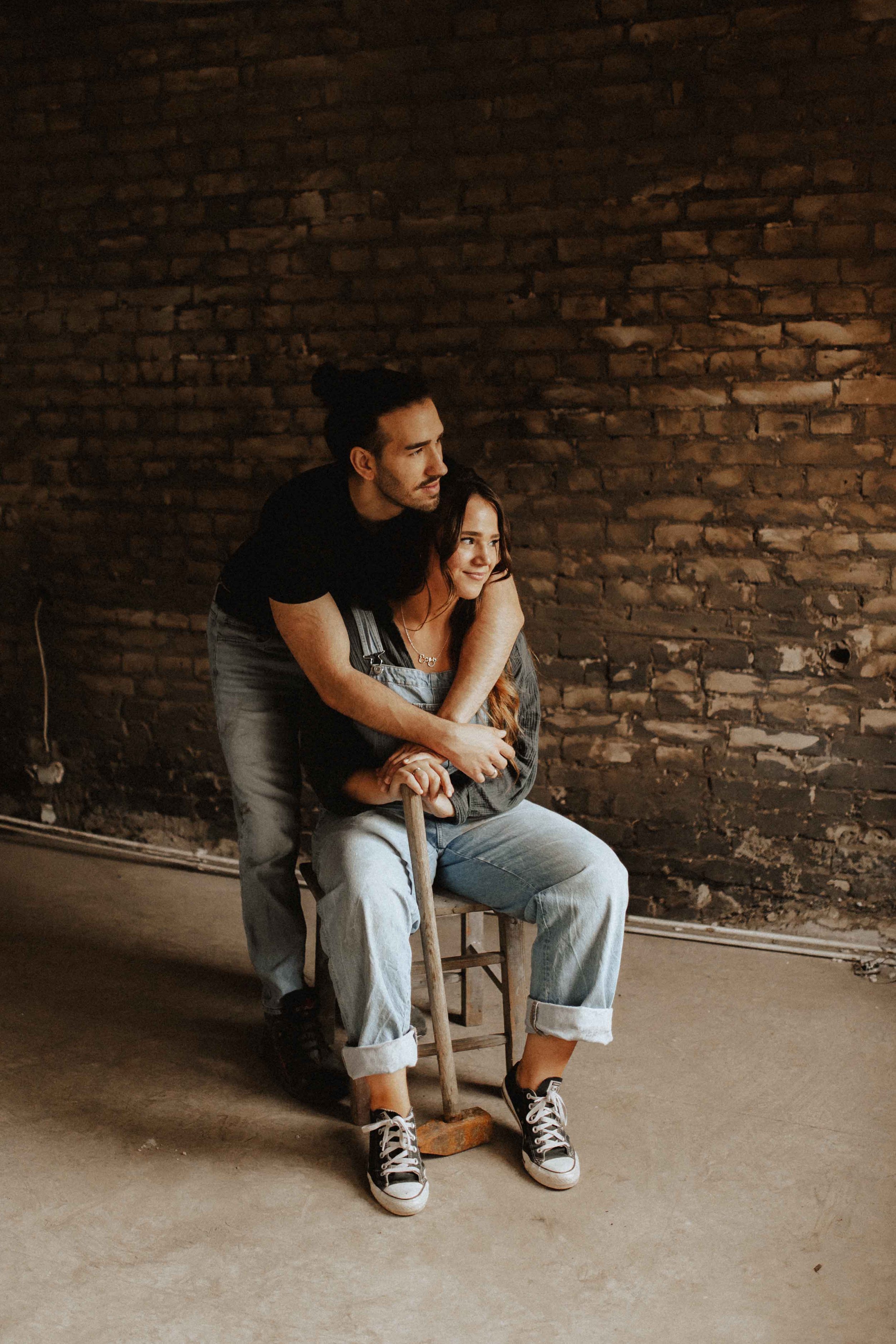 Paarshooting in Uelzen – Mann umarmt seine Partnerin von hinten, während sie lachend auf einem Stuhl vor einer alten Backsteinwand sitzt.