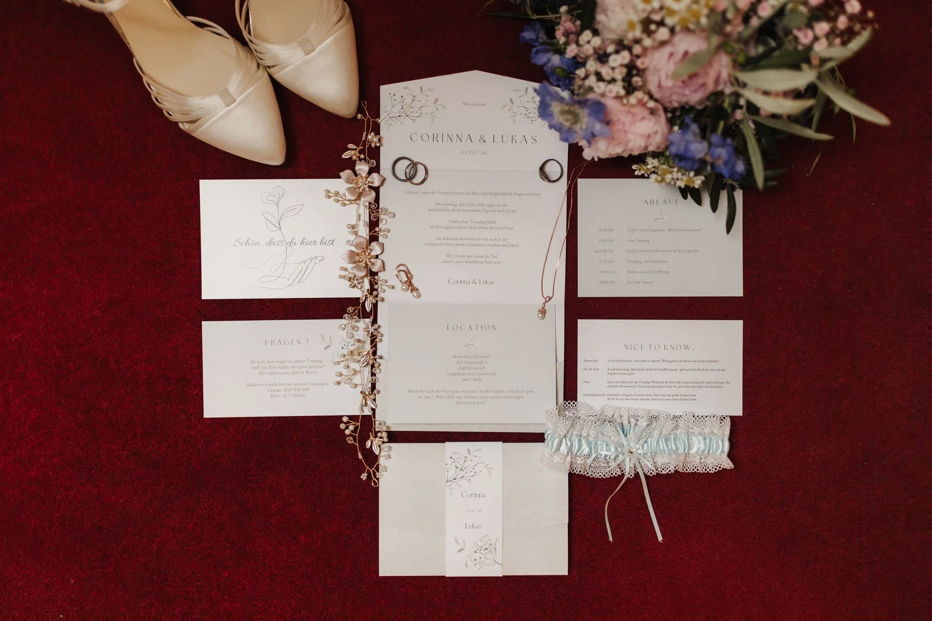 Elegantes Hochzeits-Flatlay mit Einladungskarten, Trauringen, weißen Brautschuhen, Brautschmuck und buntem Brautstrauß auf dunkelrotem Hintergrund.