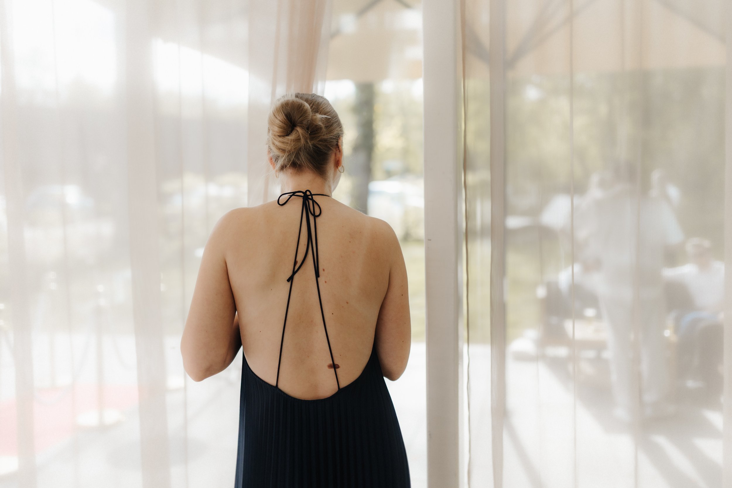 Hochzeitsgast in elegantem schwarzem, rückenfreiem Kleid mit Schleifenbindung steht am Fenster einer Hochzeitslocation in Lüneburg.