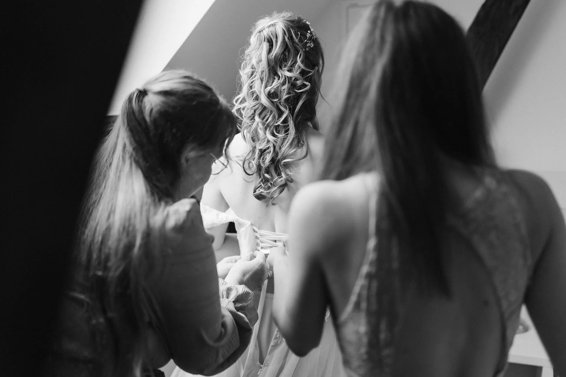 Schwarz-Weiß-Aufnahme vom Getting Ready der Braut, während zwei Frauen ihr beim Anziehen und Schnüren des Brautkleides helfen – natürliche Hochzeitsreportage im Heide Hotel Reinstorf bei Lüneburg.