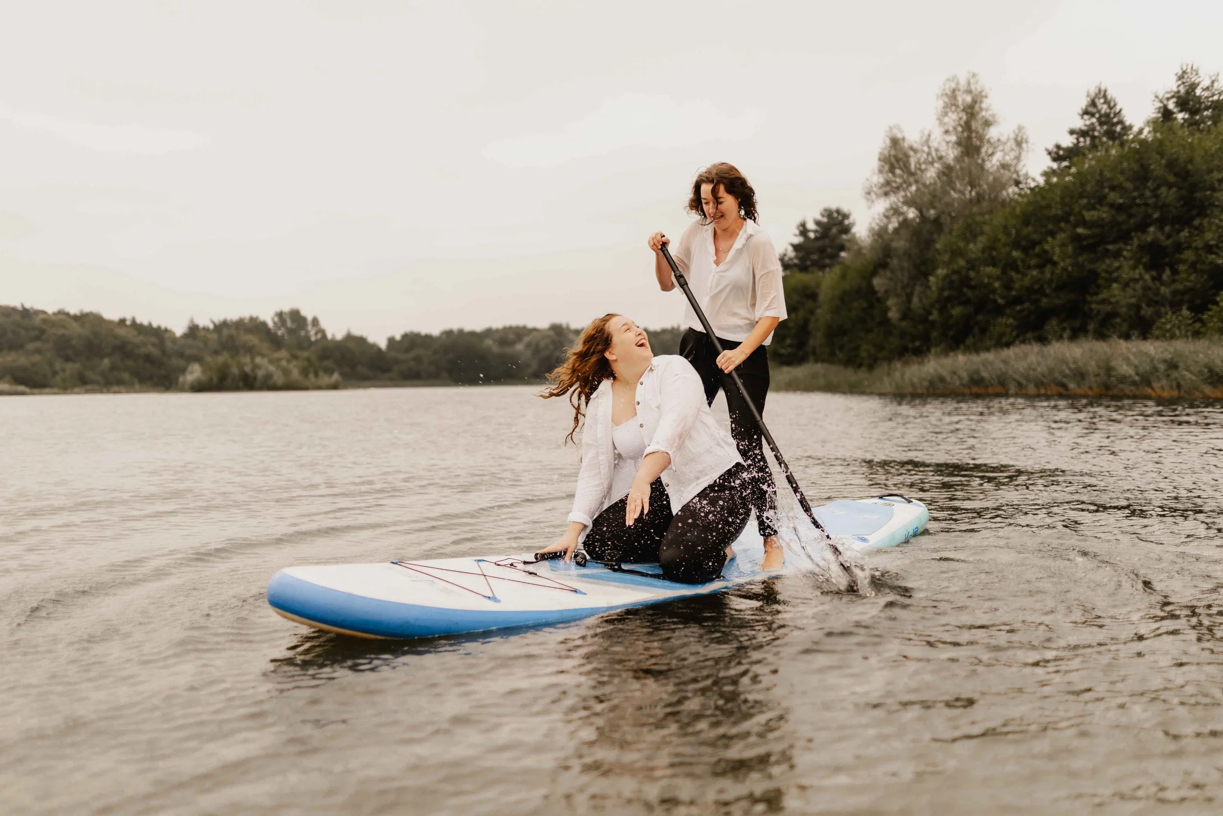Eine Frau steht paddelnd auf dem SUP, während ihre Partnerin vorne lachend kniet – Paarshooting auf dem Wasser