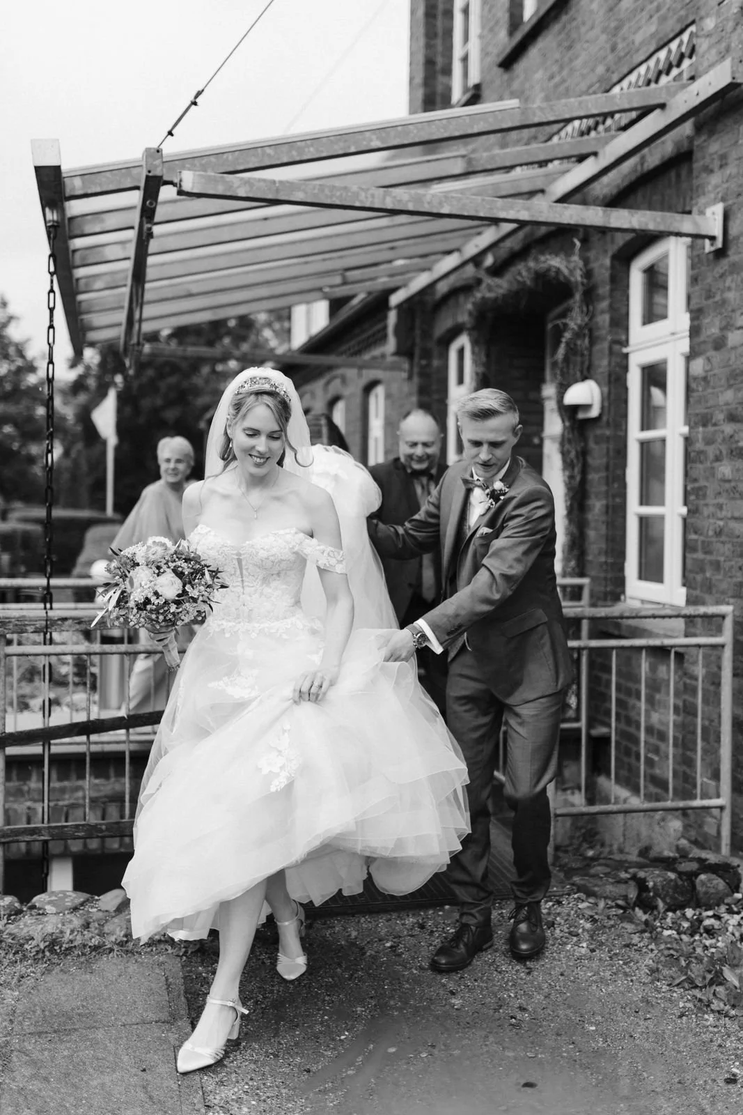 Brautpaar verlässt nach der freien Trauung das Heide Hotel Reinstorf bei Lüneburg. Der Bräutigam hilft seiner Braut im Hochzeitskleid über die Stufe – ein natürlicher Moment der Hochzeitsreportage in Schwarz-Weiß.