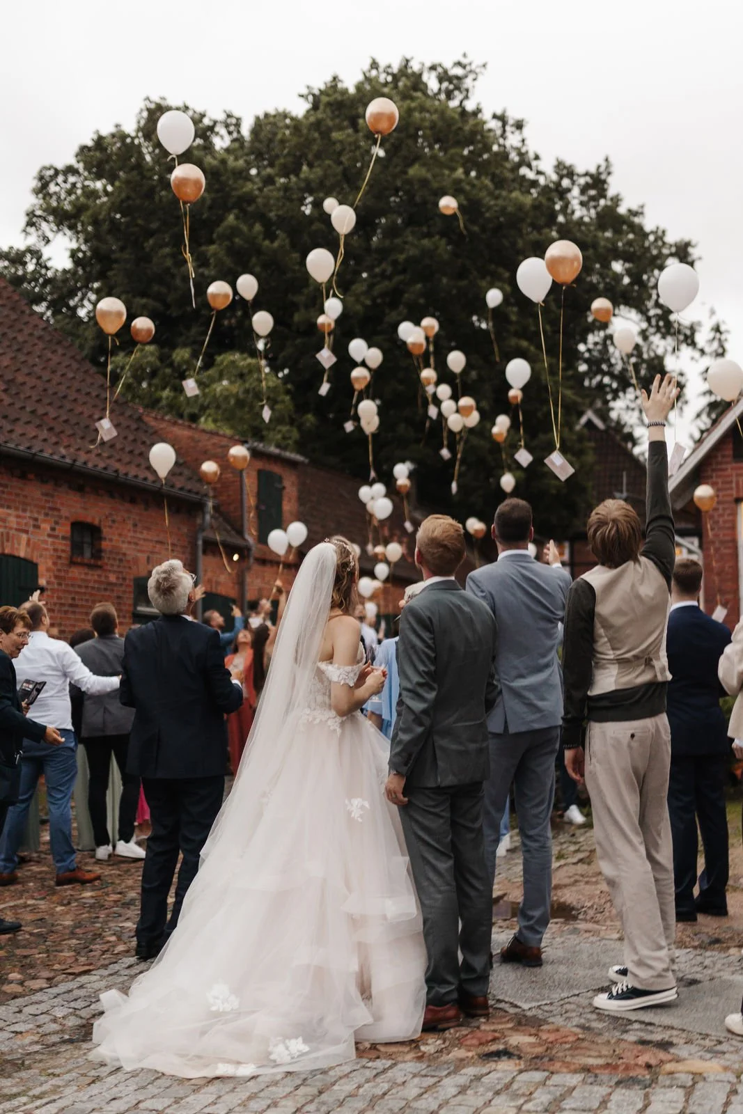 Hochzeitsgesellschaft beim gemeinsamen Luftballonsteigenlassen im Innenhof des Heide Hotels Reinstorf bei Lüneburg. Das Brautpaar und seine Gäste beobachten, wie zahlreiche weiß-goldene Luftballons über den historischen Backsteingebäuden in den Himme