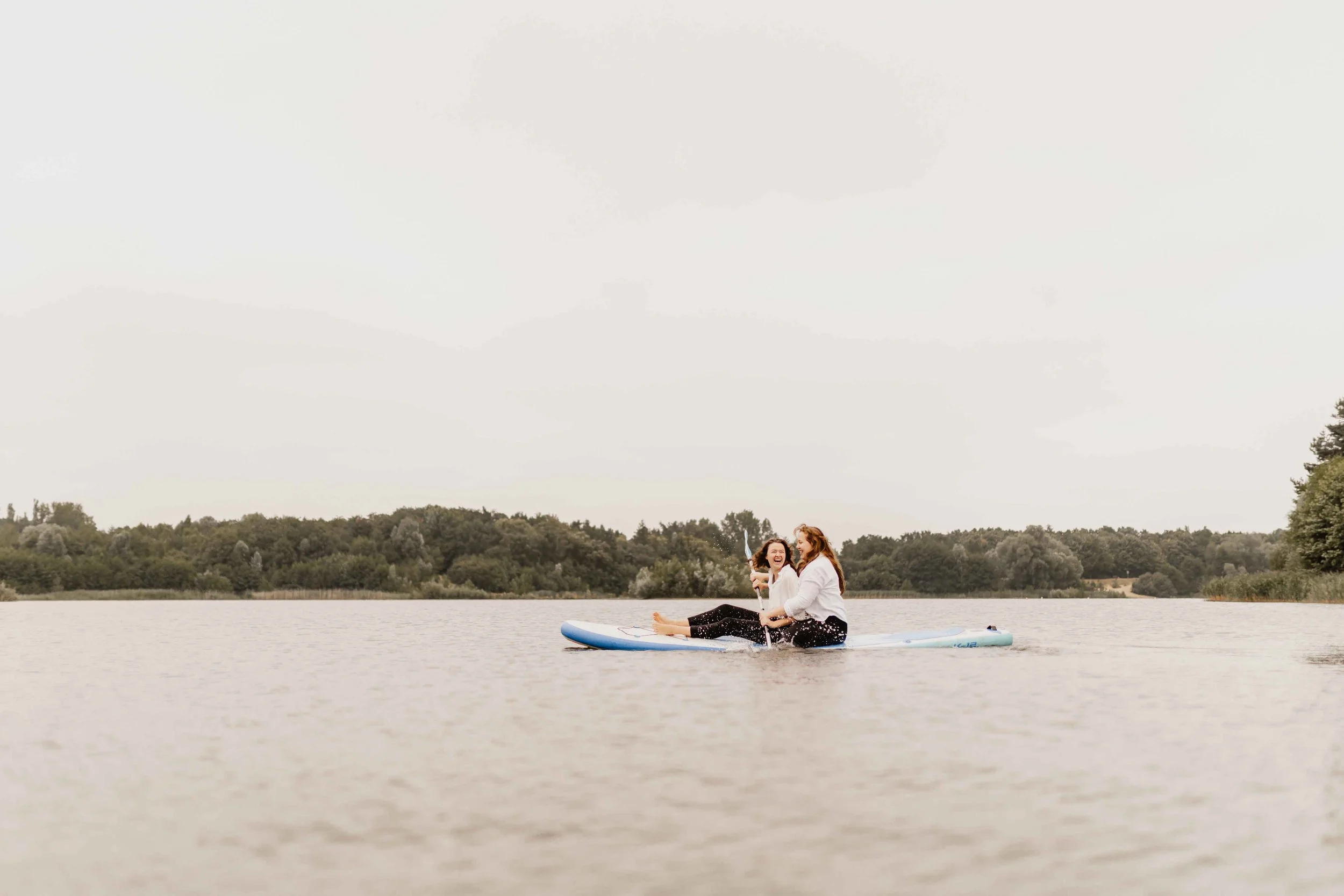 Zwei Frauen sitzen gemeinsam auf einem SUP-Board auf dem See, umgeben von Wasser und Natur