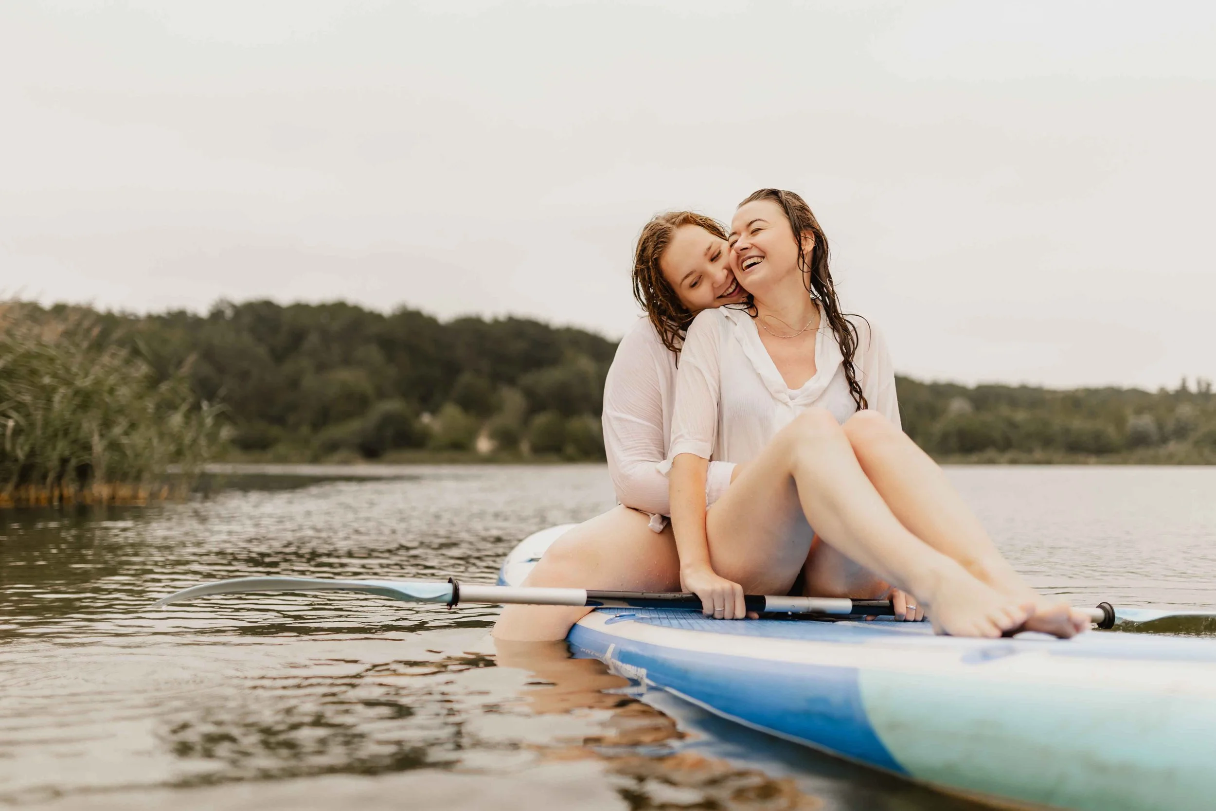 Zwei Frauen sitzen eng umschlungen auf einem SUP-Board auf dem See und genießen die gemeinsame Zeit