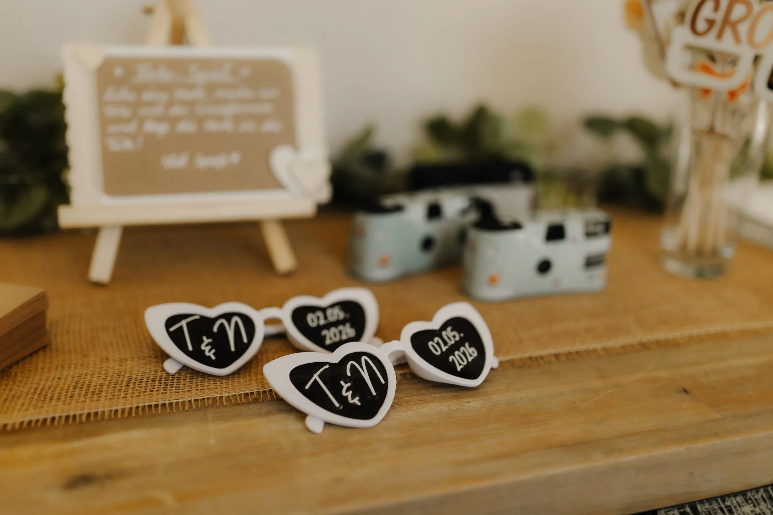 Herzförmige Hochzeitsbrillen mit den Initialen von Tina und Niklas sowie dem Hochzeitsdatum neben Sofortbildkameras als kreative Fotoaccessoires für die Hochzeitsgäste.