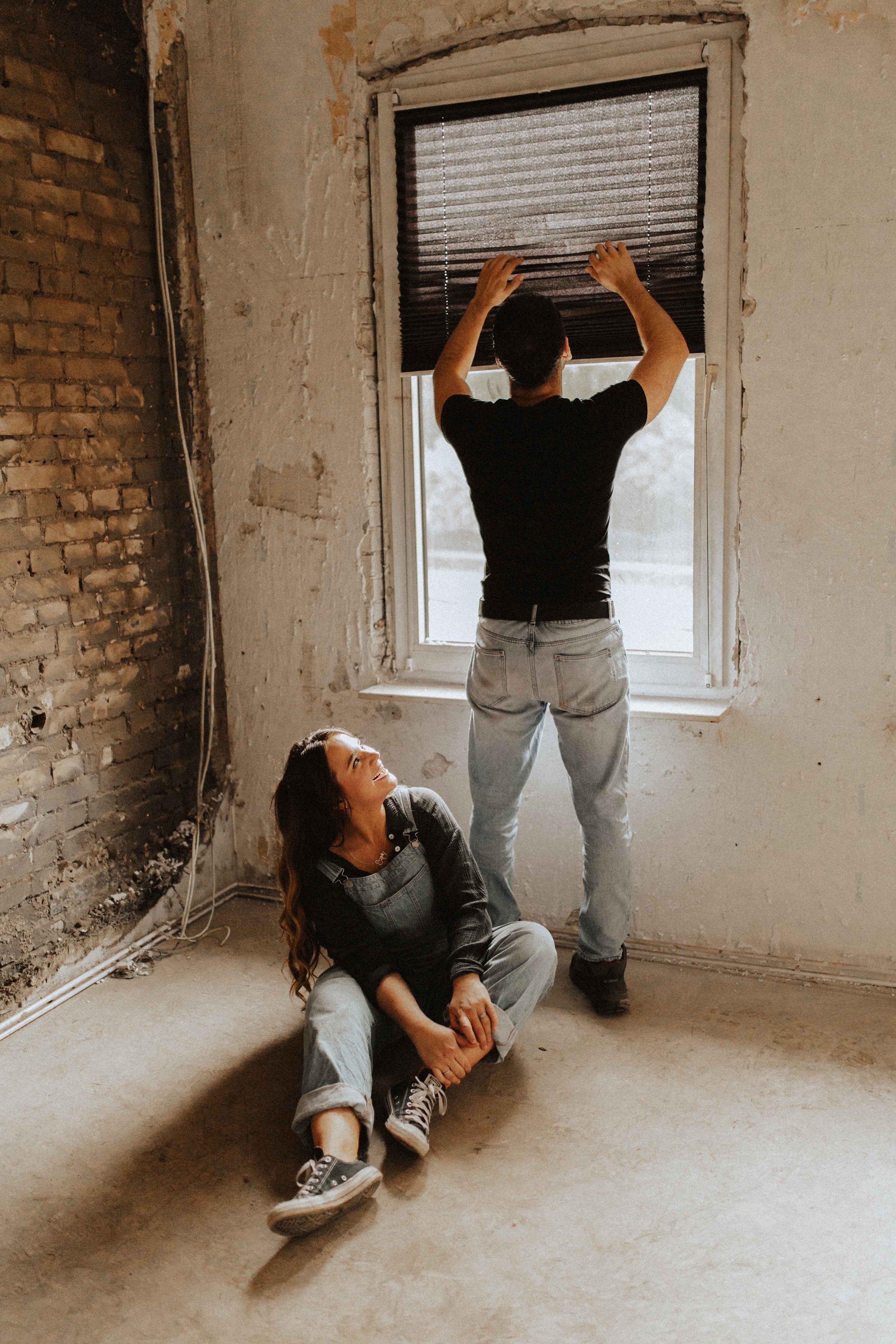 Authentisches Paarshooting in Lüneburg – Mann öffnet die Jalousie, während die Frau lachend am Boden sitzt und zu ihm aufschaut.