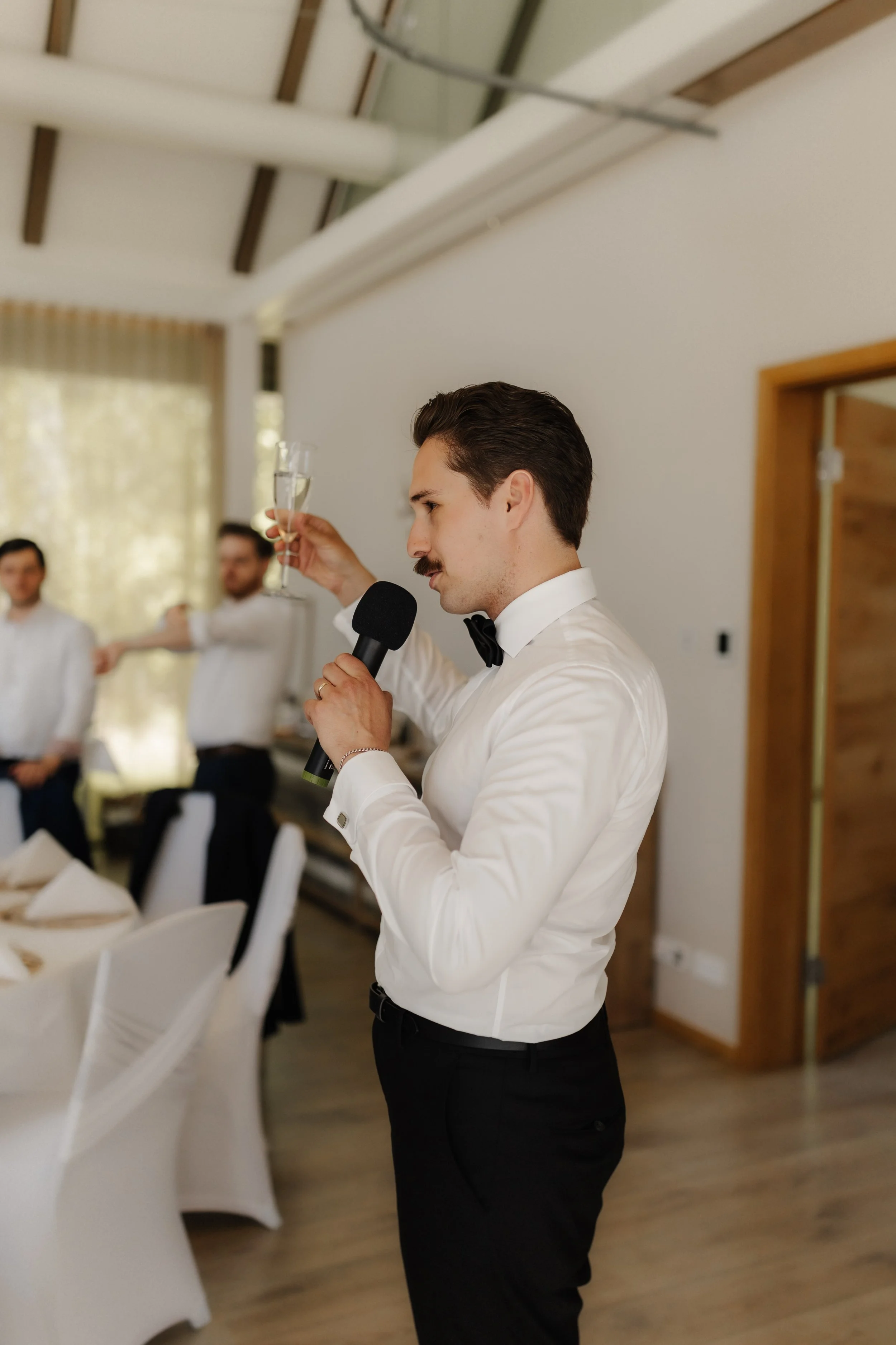 Bräutigam Niklas hält während seiner Hochzeitsrede ein Sektglas hoch und spricht mit Mikrofon zu den Gästen im Forsthaus Rote Schleuse in Lüneburg.