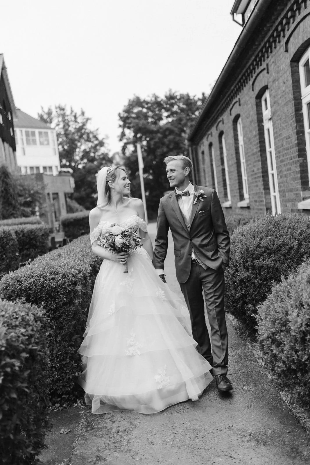 Brautpaar spaziert Hand in Hand und lächelnd am Heide Hotel Reinstorf bei Lüneburg entlang – natürliche Schwarz-Weiß-Hochzeitsfotografie voller Liebe und Leichtigkeit.