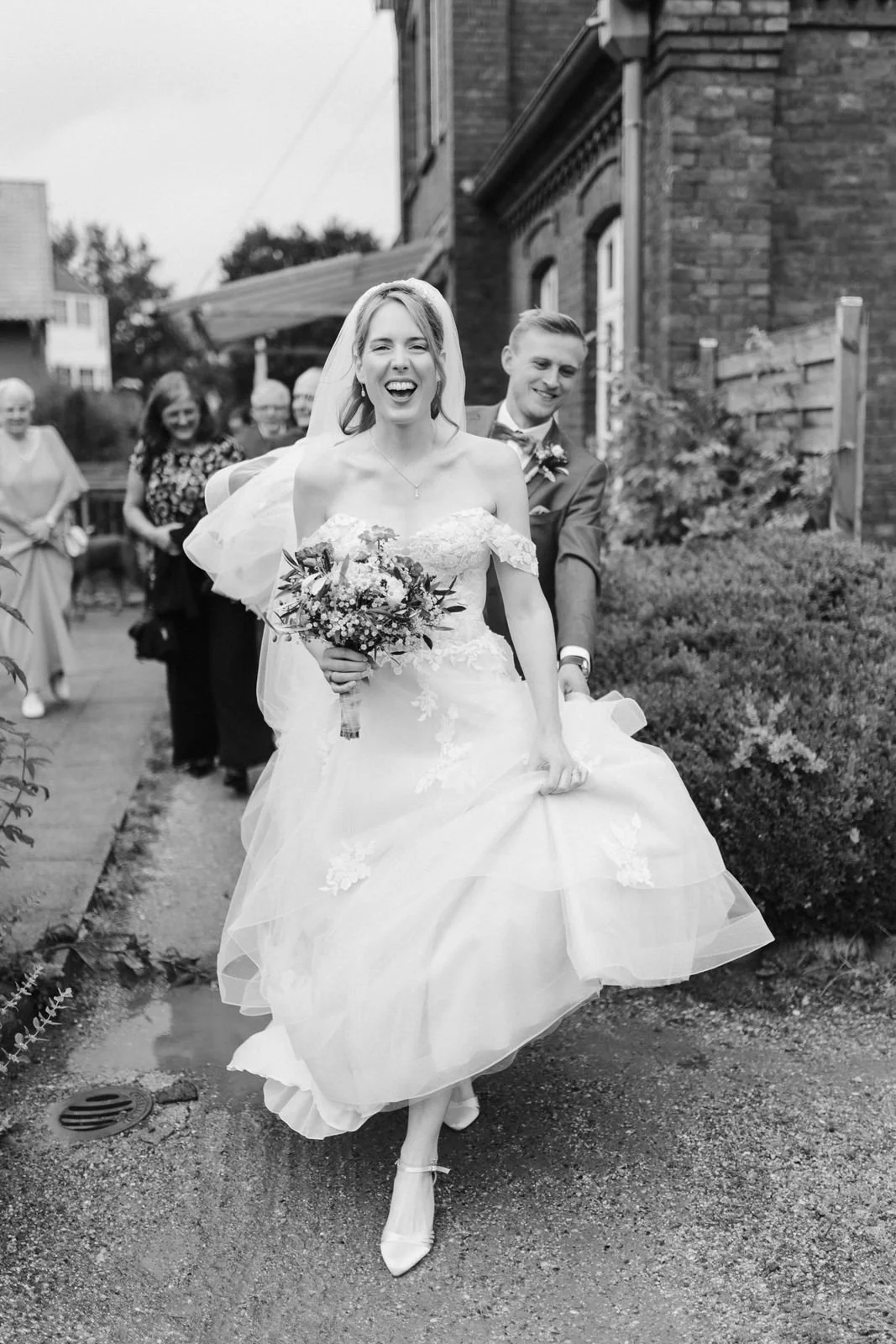 Strahlende Braut mit Brautstrauß und ihrem Bräutigam beim fröhlichen Spaziergang nach der Hochzeit im Heide Hotel Reinstorf bei Lüneburg – authentische Hochzeitsfotografie in Schwarz-Weiß.