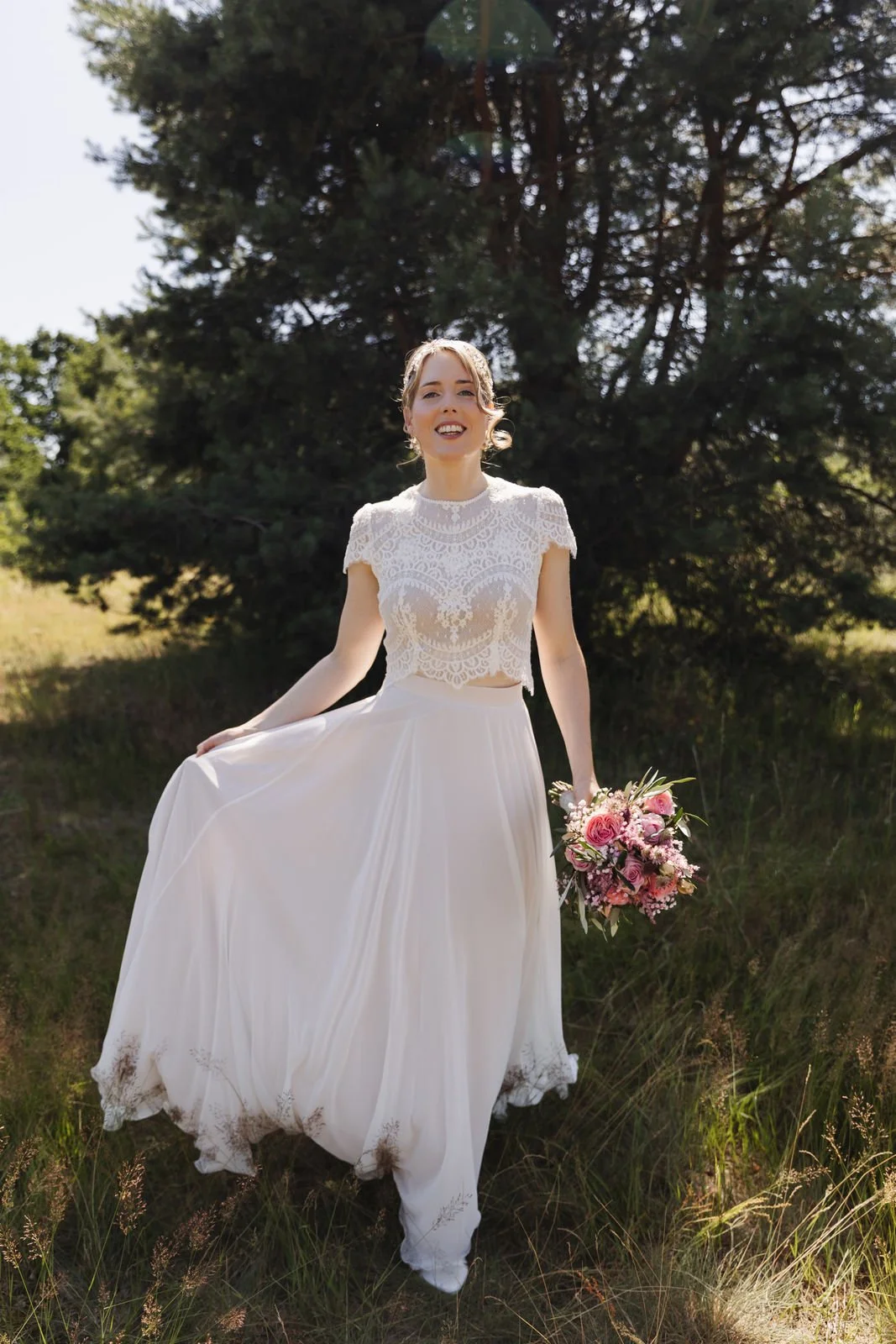 Strahlende Braut im weißen Hochzeitskleid mit Spitzenoberteil und rosa Brautstrauß. Beim Brautporträt nahe dem Heide Hotel Reinstorf hält sie lächelnd ihren fließenden Rock.
