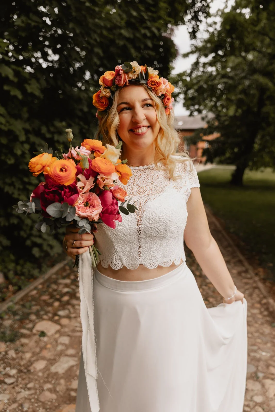 Strahlende Braut mit Blumenkranz und Brautstrauß blickt in die Kamera auf einem Weg in Lüneburg