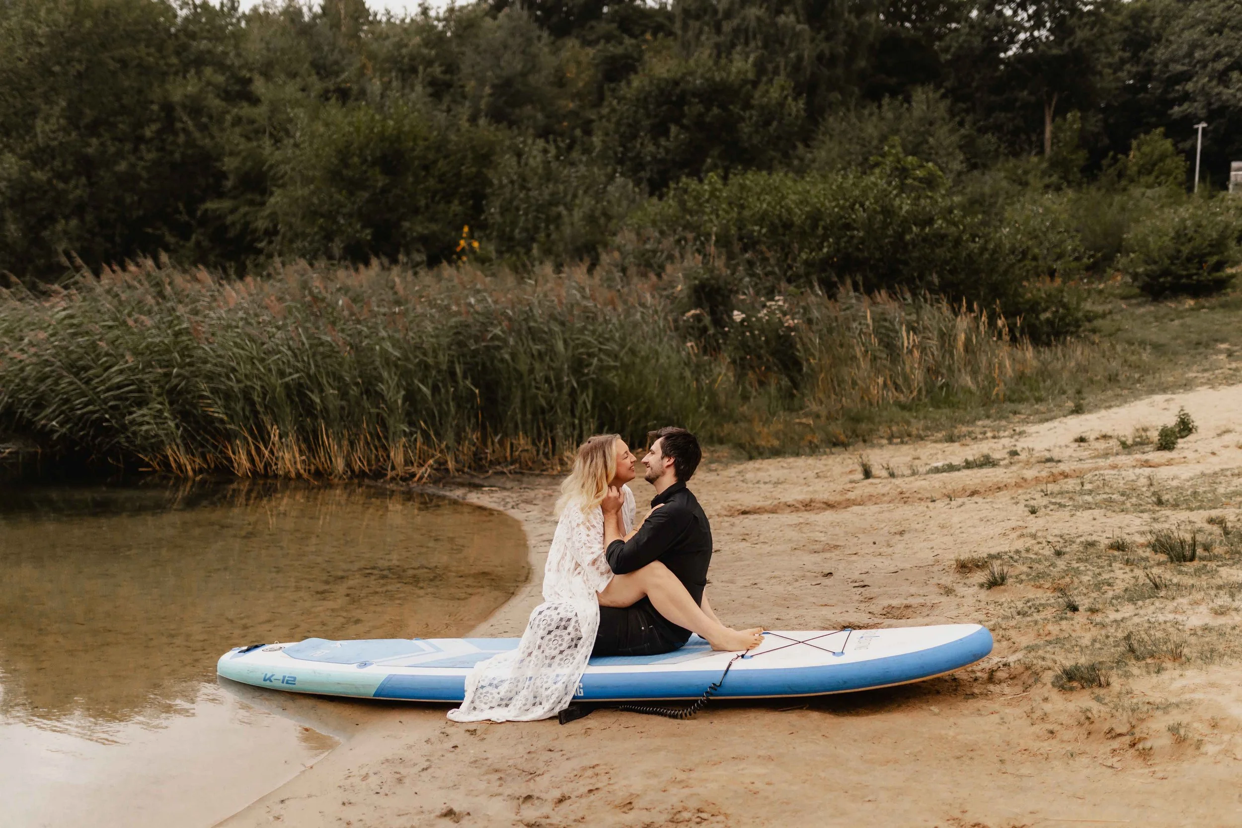 Paar sitzt eng umschlungen auf einem SUP am Ufer und schaut sich verliebt an