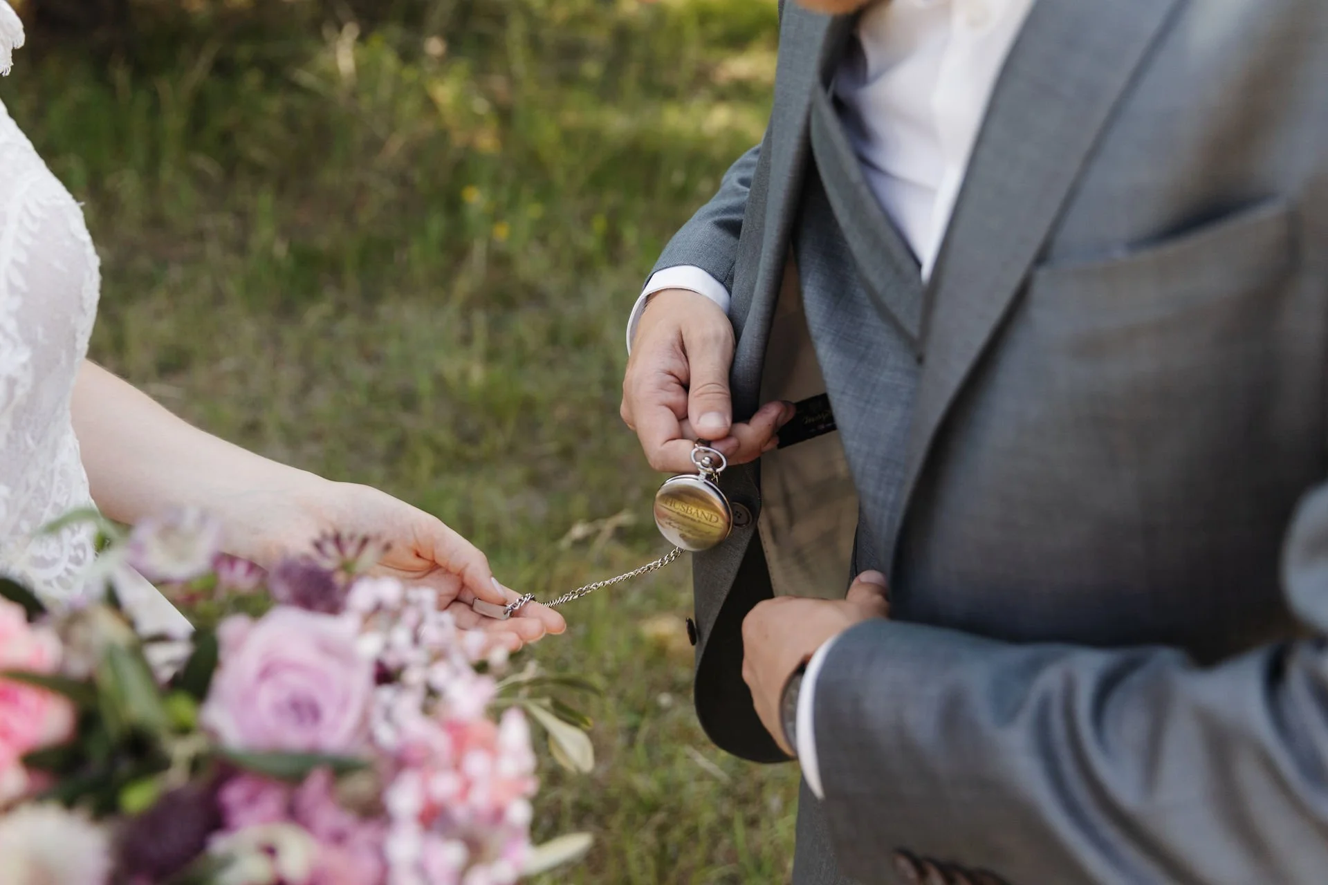 Detailaufnahme eines Brautpaares beim Hochzeitsshooting. Der Bräutigam hält eine goldene Taschenuhr, während die Braut die Kette berührt. Im Vordergrund ein rosa Brautstrauß.