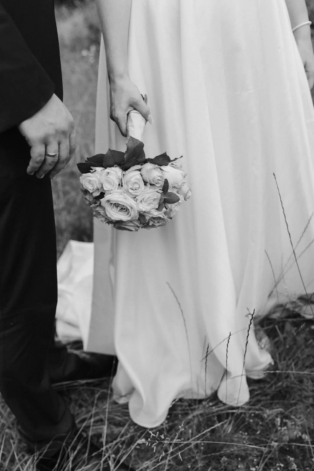 Detailaufnahme eines Brautpaares Hand in Hand, während die Braut ihren Brautstrauß neben dem fließenden Hochzeitskleid trägt – Schwarz-Weiß-Fotografie