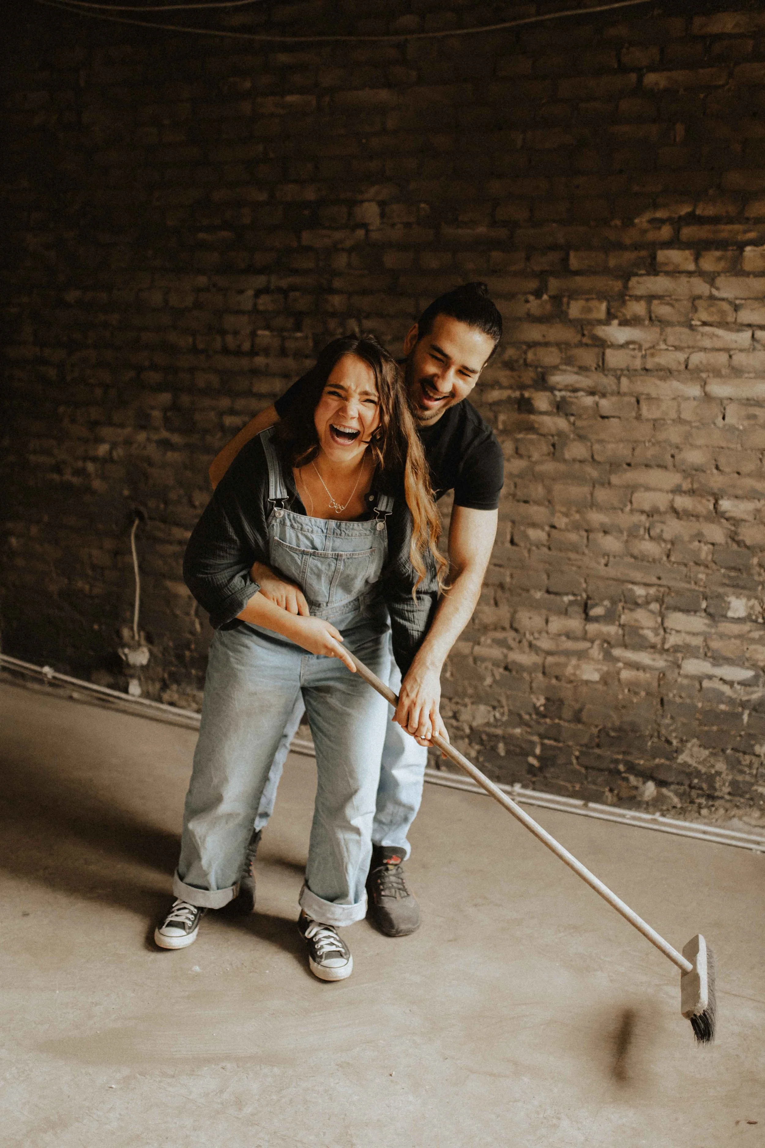Paarshooting auf Baustelle – lachendes Paar fegt gemeinsam den Boden vor Backsteinwand in Lüneburg.