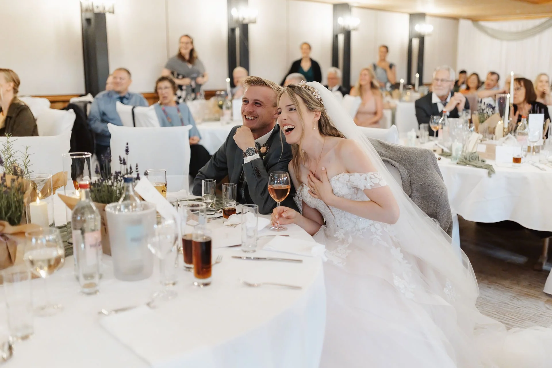 Hochzeitsfotografie vom Brautpaar, die bei ihrer Hochzeitsfeier im Heide Hotel Reinstorf bei Lüneburg gemeinsam herzlich lachen, während die Gäste im Hintergrund zuschauen.