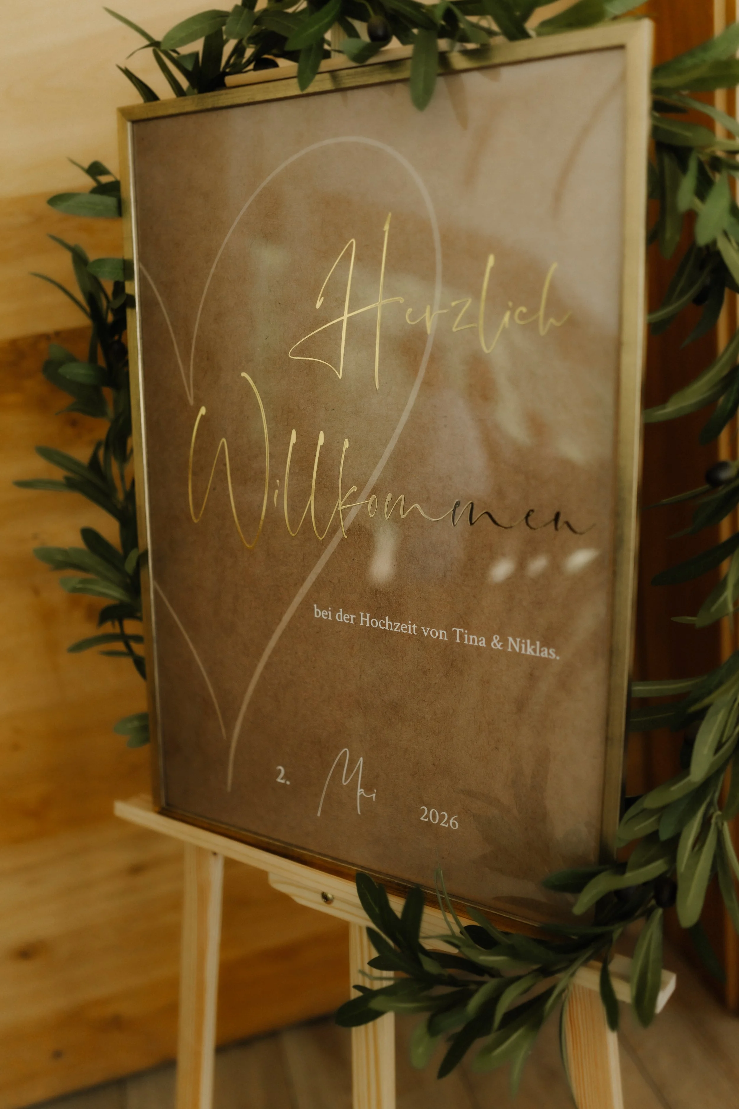 Elegantes Willkommensschild zur Hochzeit von Tina und Niklas mit goldener Schrift, Herzmotiv und grüner Pflanzendekoration.
