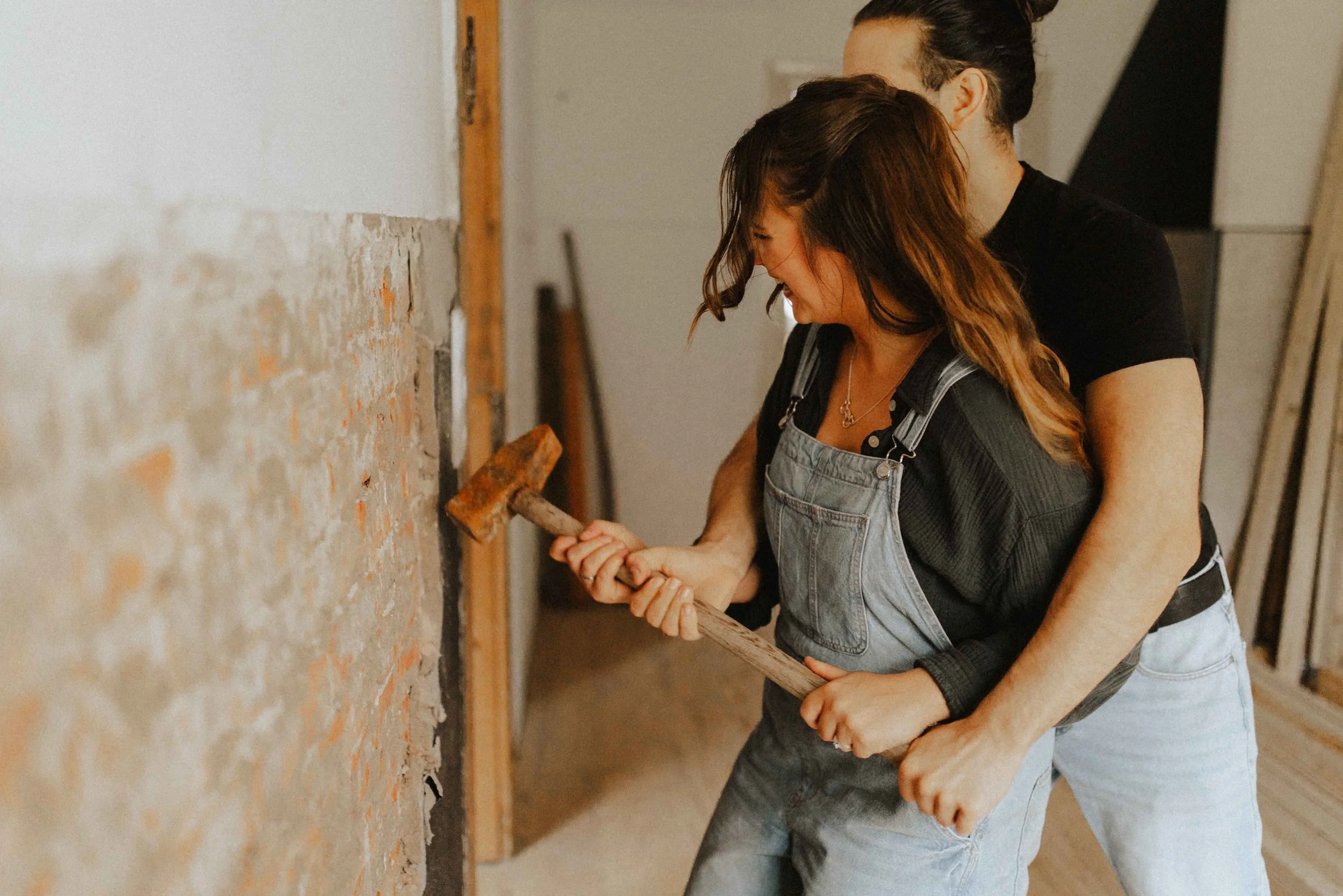 Baustellenshooting in Lüneburg – verliebtes Paar in Arbeitskleidung, Mann umarmt Frau von hinten, während sie gemeinsam einen Hammer hält.