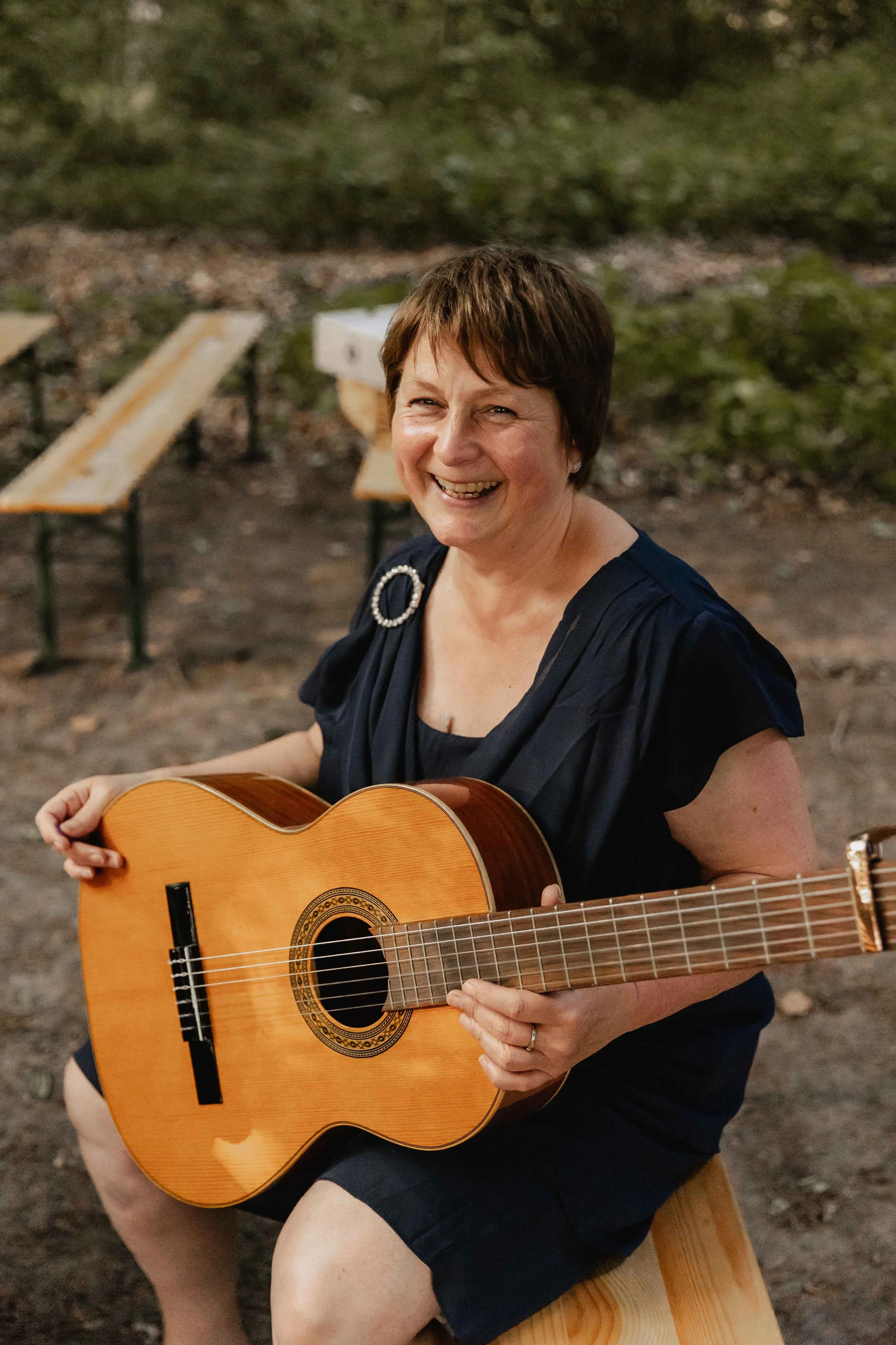 Musikerin mit Gitarre lächelt herzlich auf Holzbänken im Wald