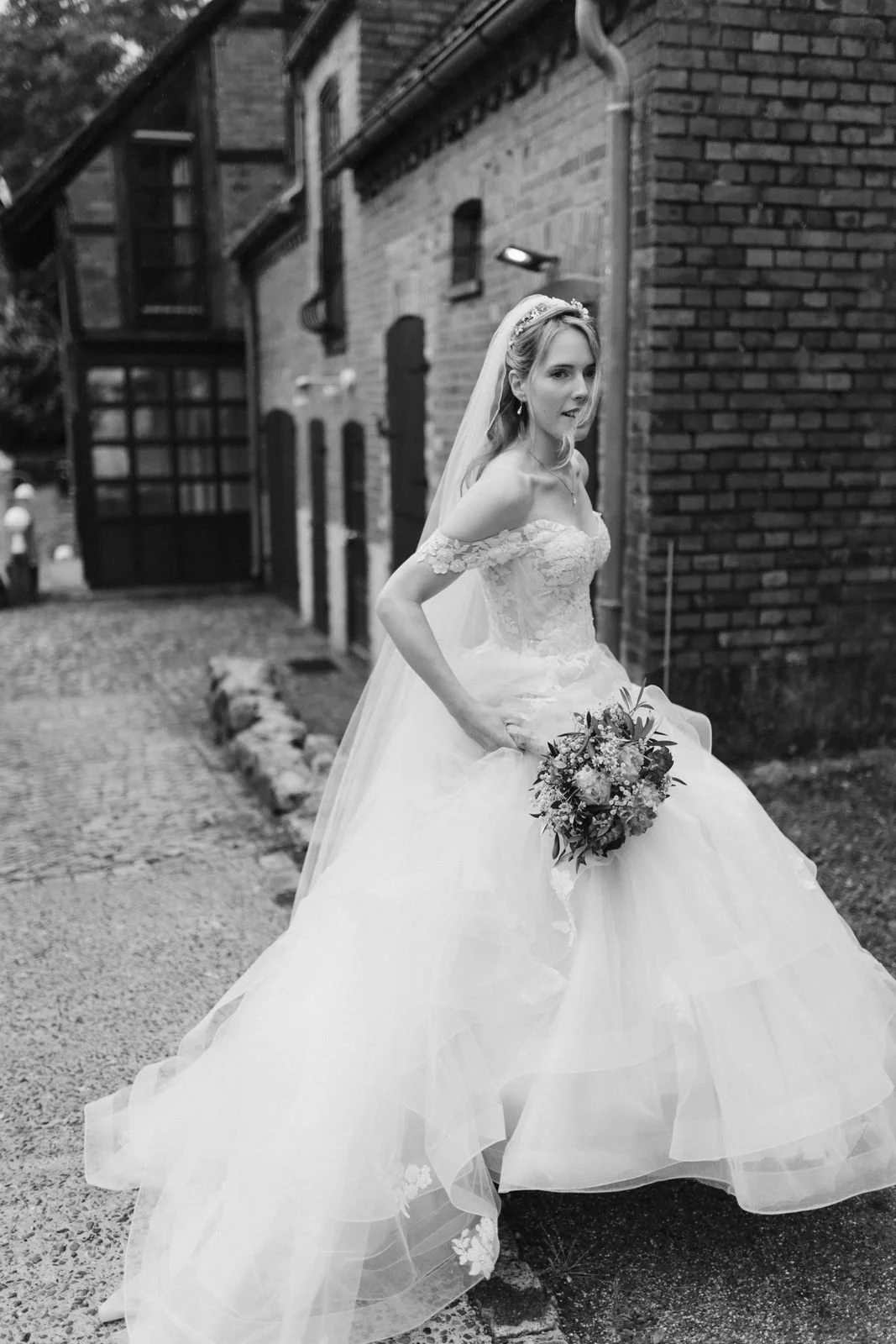 Stimmungsvolle Schwarz-Weiß-Aufnahme einer Braut im romantischen Brautkleid mit langer Schleppe, Schleier und Brautstrauß beim Hochzeitsshooting vor der historischen Backsteinfassade des Heide Hotels Reinstorf bei Lüneburg.