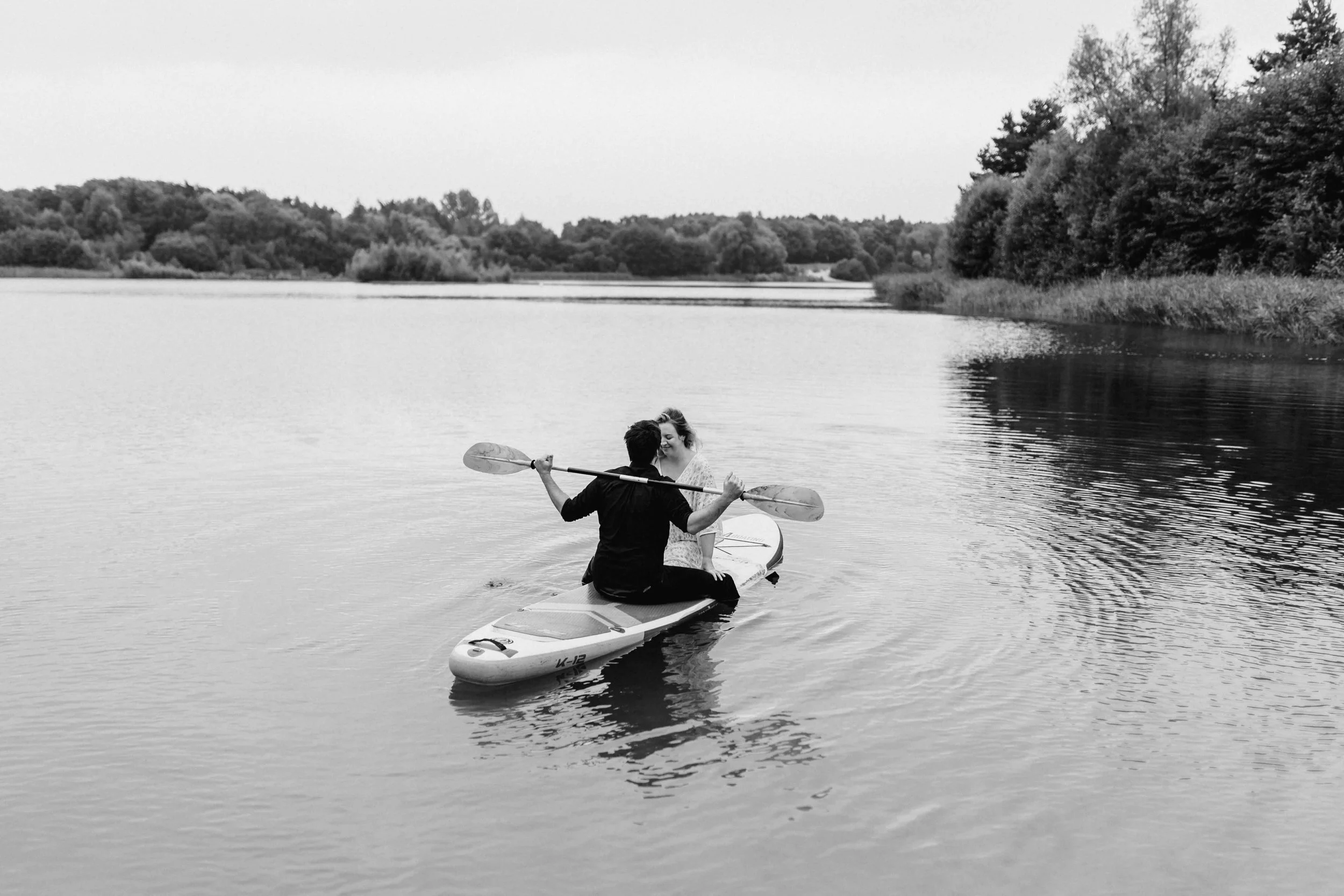 Paar sitzt gemeinsam auf SUP-Board am See, er hält ein Paddel während sie sich zärtlich anlehnen, Schwarz-Weiß-Fotografie