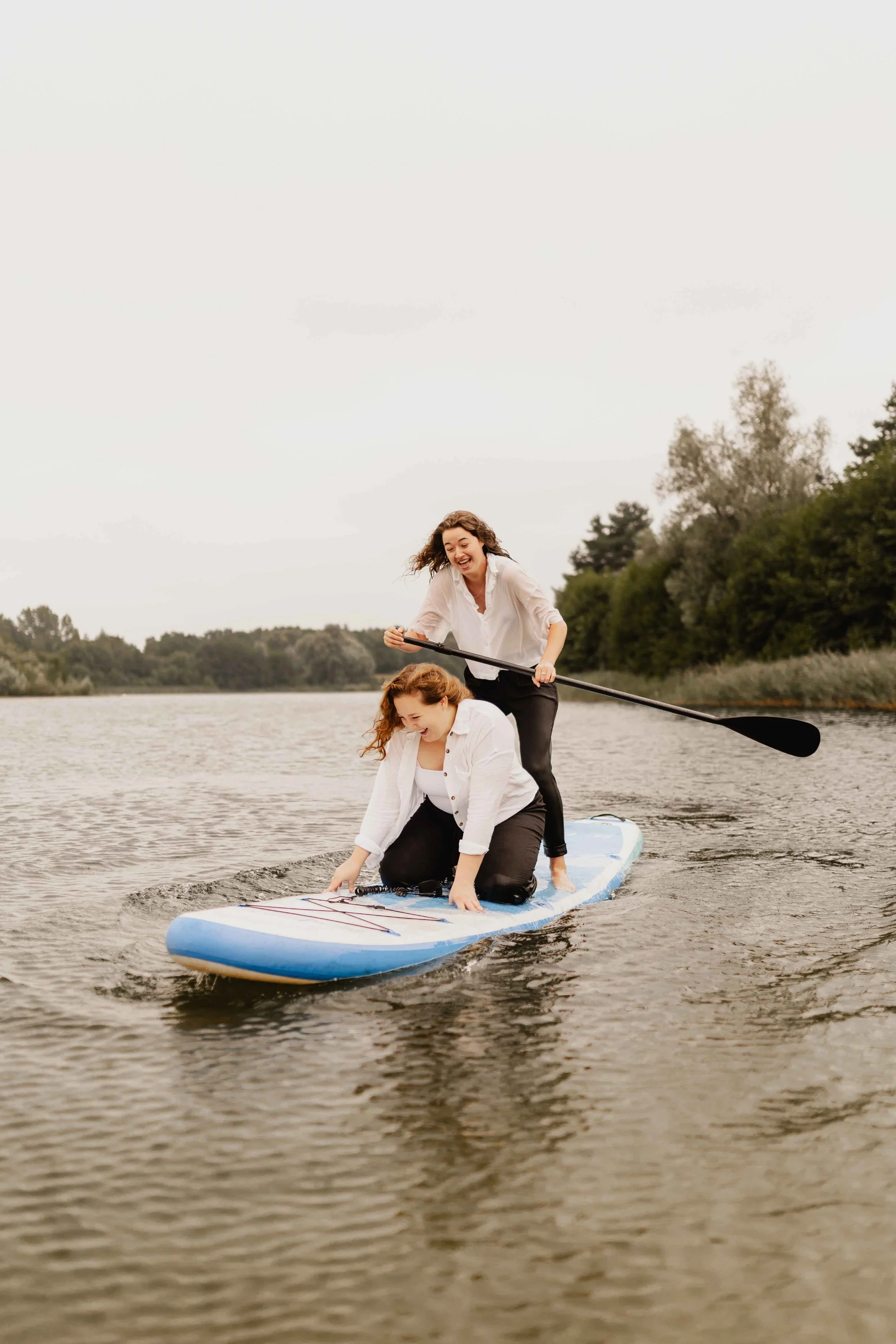 Eine Frau steht mit Paddel auf einem SUP, während ihre Partnerin lachend davor sitzt – Paarshooting auf dem Wasser