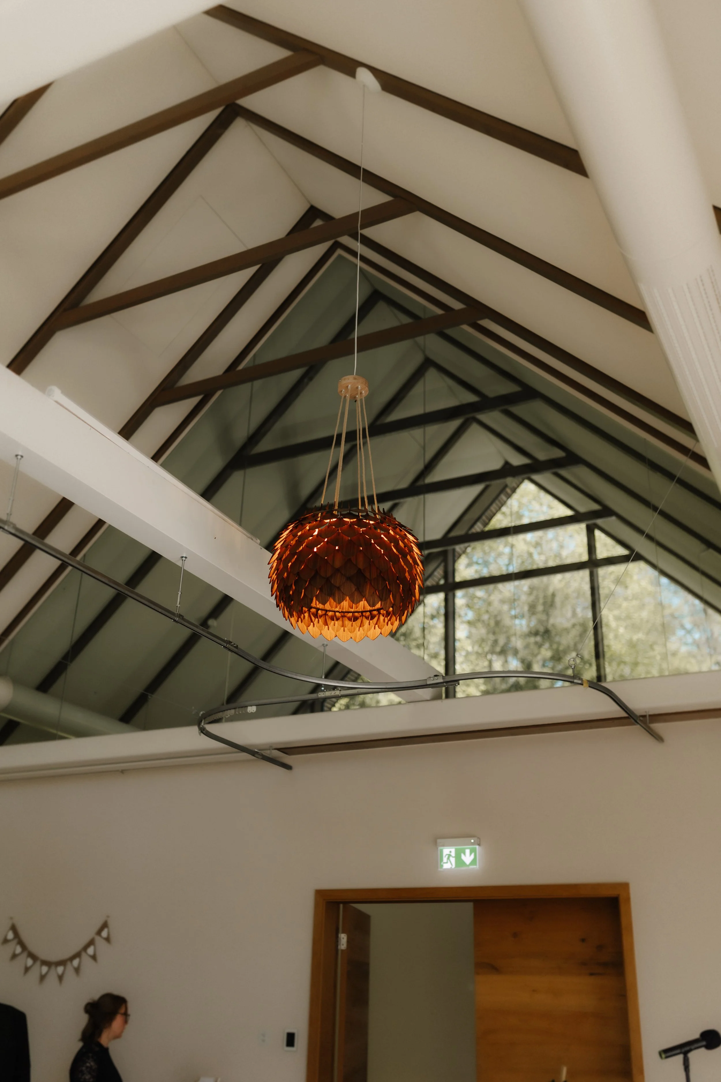 Innenansicht einer modernen Hochzeitslocation mit hohen Glasfronten, sichtbaren Holzbalken und einer außergewöhnlichen Pendelleuchte unter dem Spitzdach.