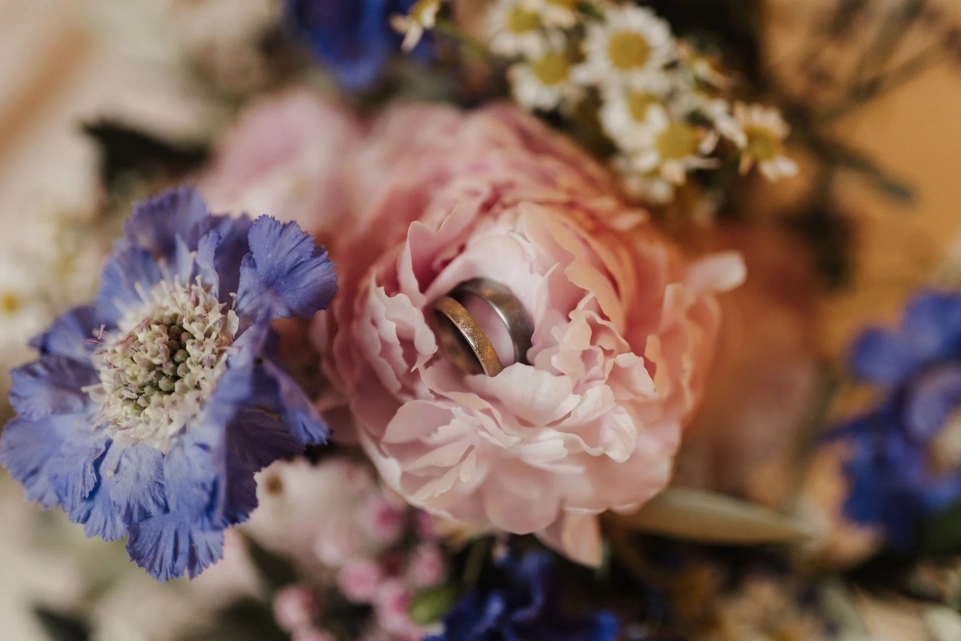 Nahaufnahme zweier Eheringe in einer rosafarbenen Blüte, umgeben von blauen und weißen Blumen im Brautstrauß einer Hochzeit im Heide Hotel Reinstorf bei Lüneburg.