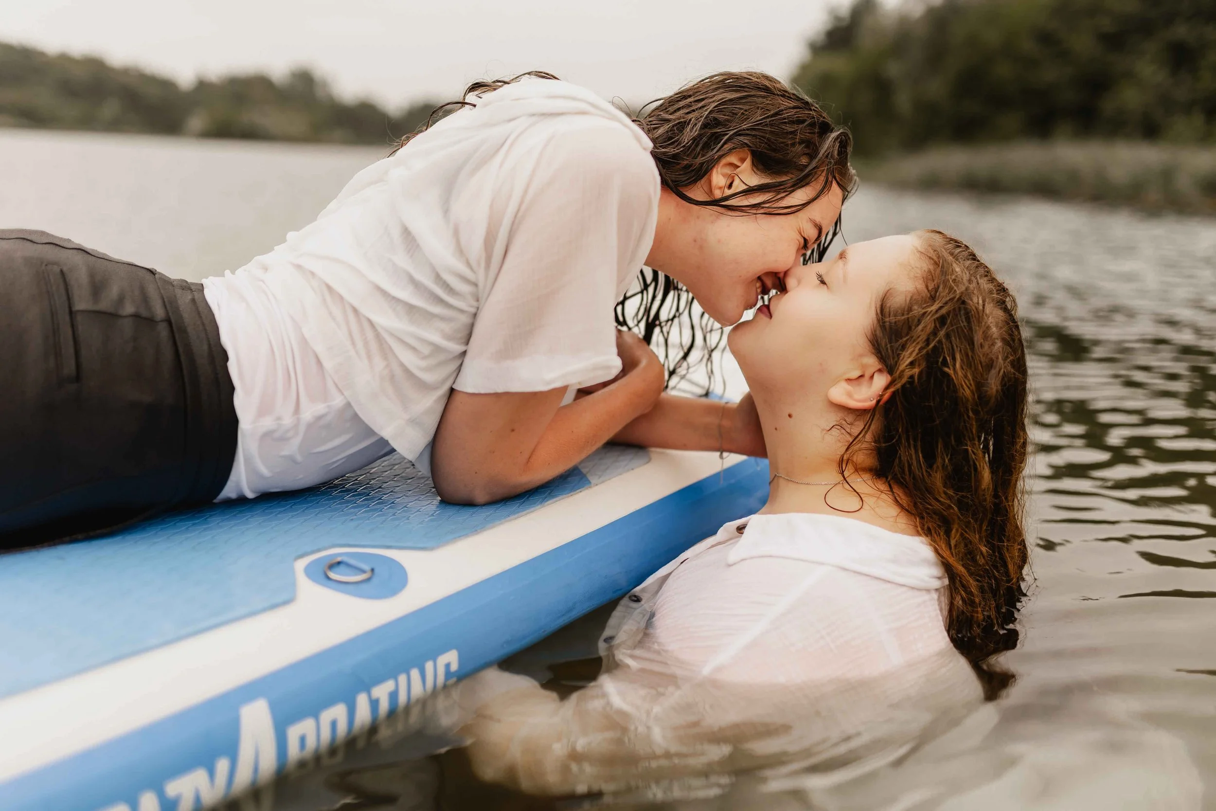 Zwei Frauen küssen sich am SUP, eine liegt darauf, die andere steht im Wasser