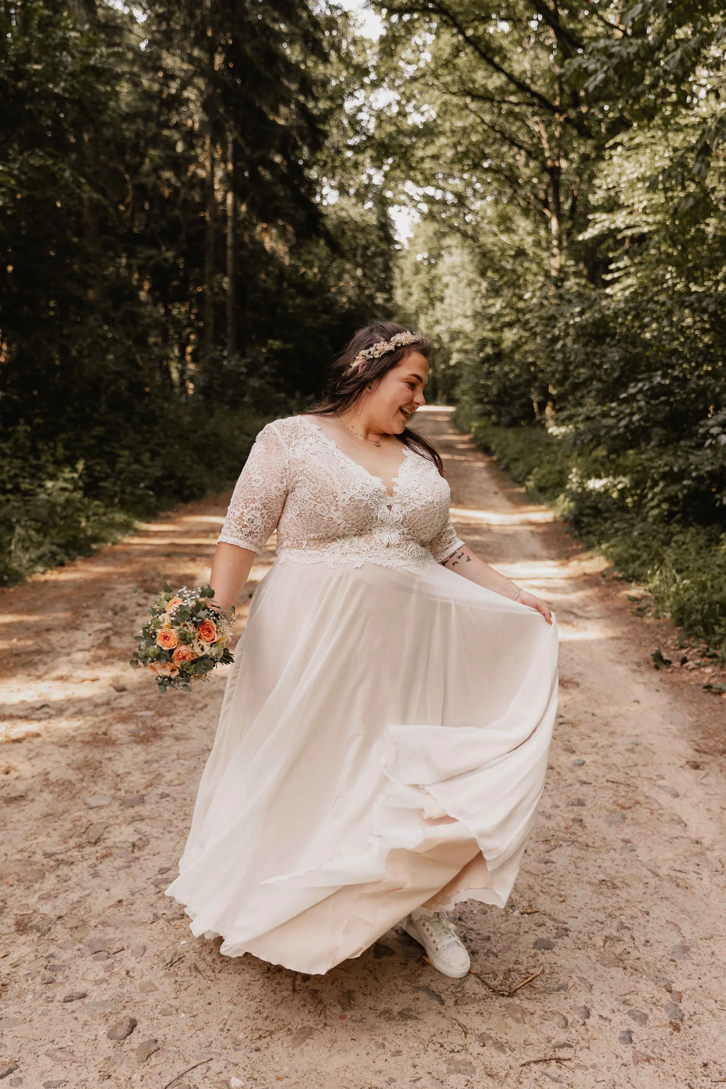 Braut dreht sich lachend im Hochzeitskleid mit Blumenstrauß auf Waldweg