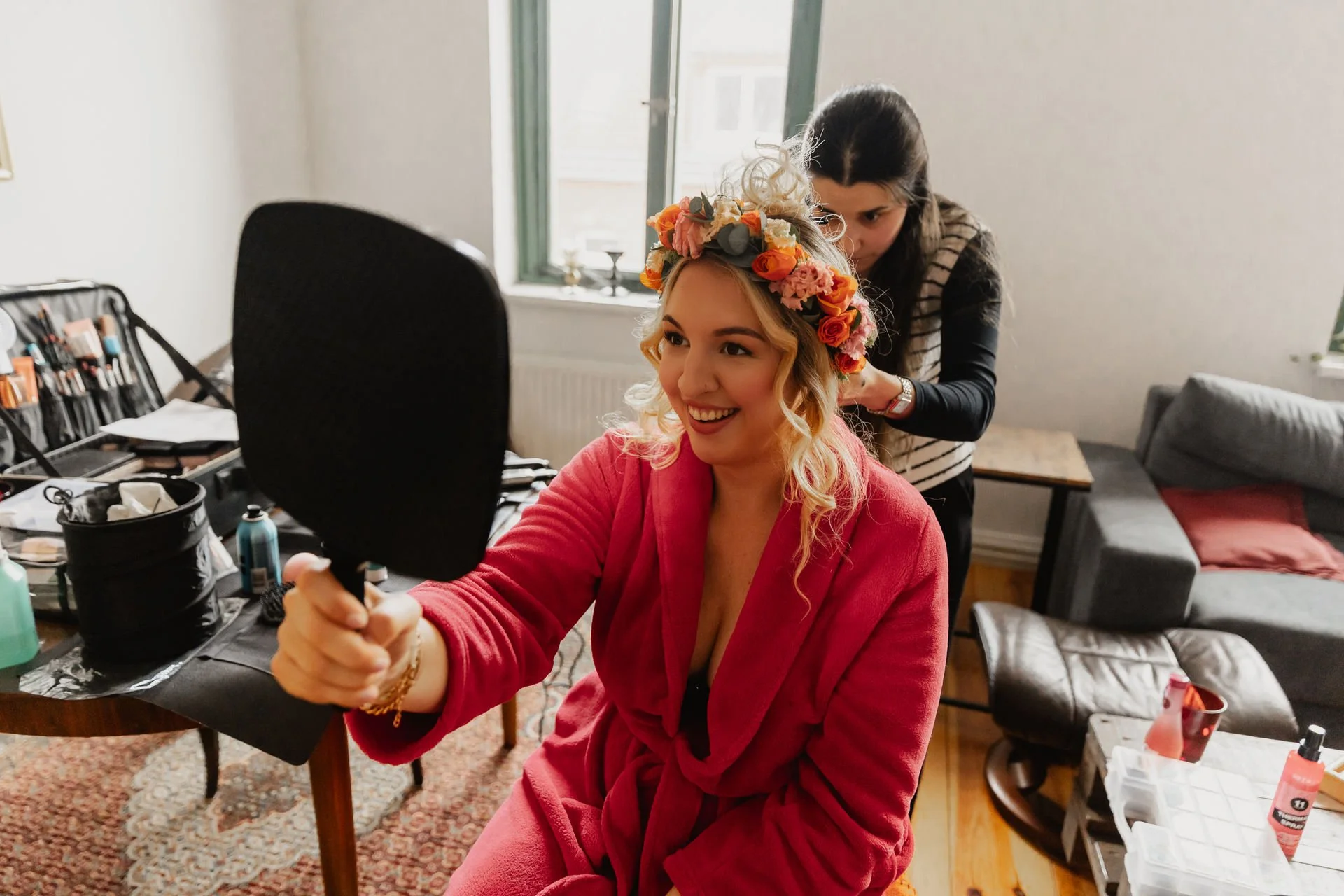Fröhliche Braut beim Getting Ready in Lüneburg mit Blumenkranz und Stylistin im Hintergrund