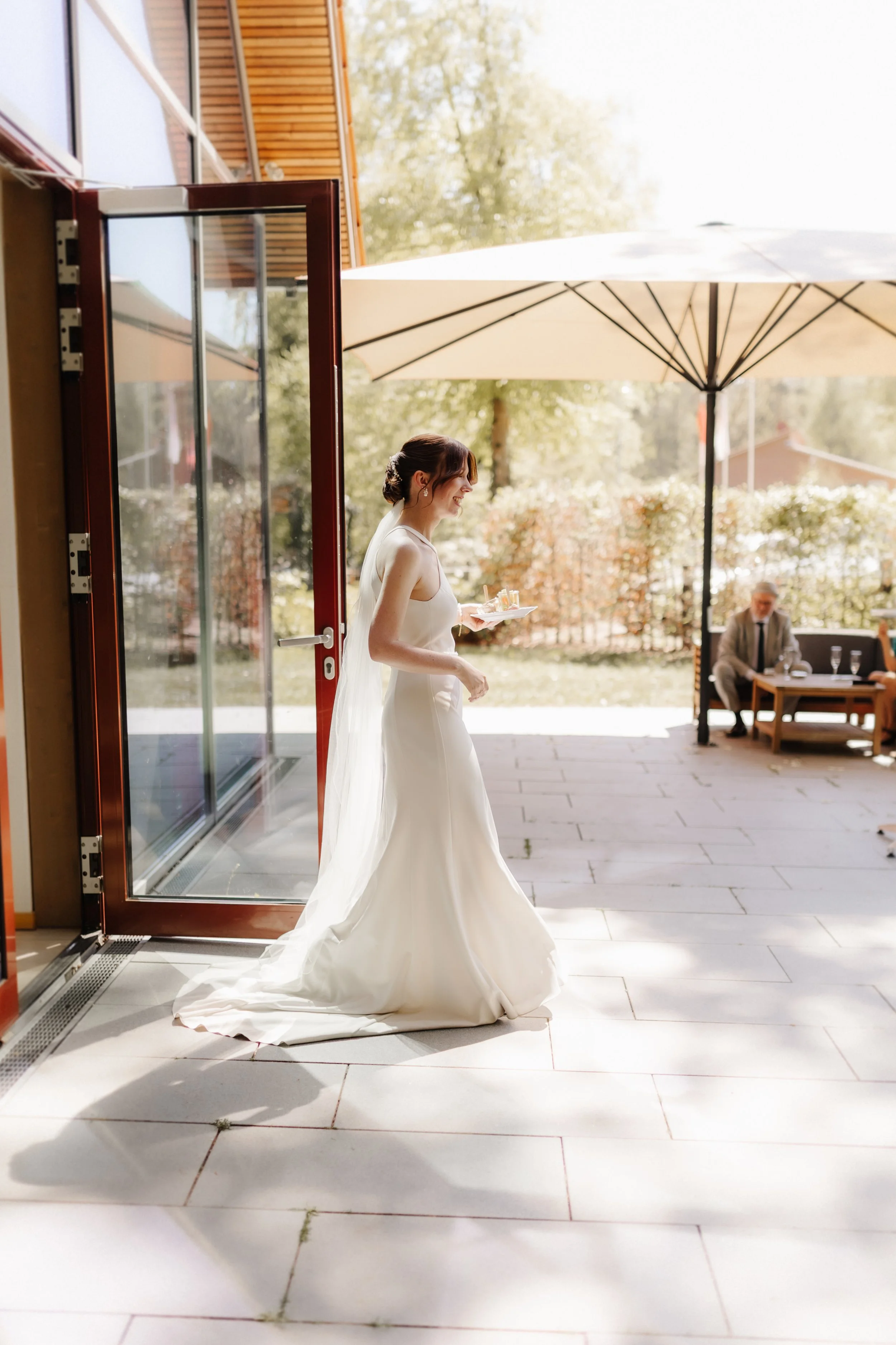 Braut im eleganten weißen Hochzeitskleid mit langer Schleppe auf der sonnigen Terrasse des Forsthauses Rote Schleuse in Lüneburg.