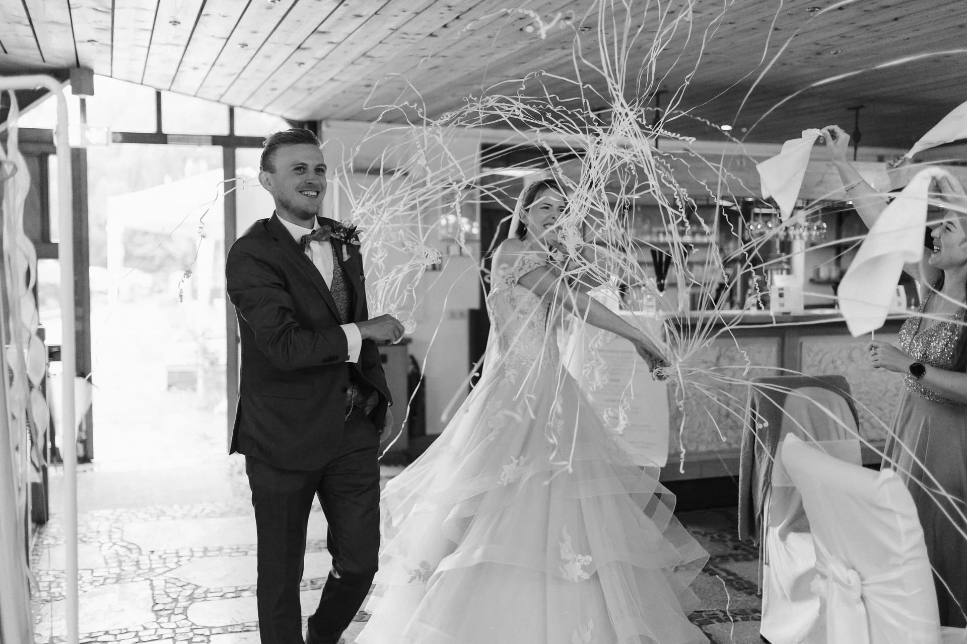 Schwarz-Weiß-Aufnahme eines lachenden Brautpaares mit fliegenden Luftschlangen beim Hochzeitsempfang im Heide Hotel Reinstorf bei Lüneburg.