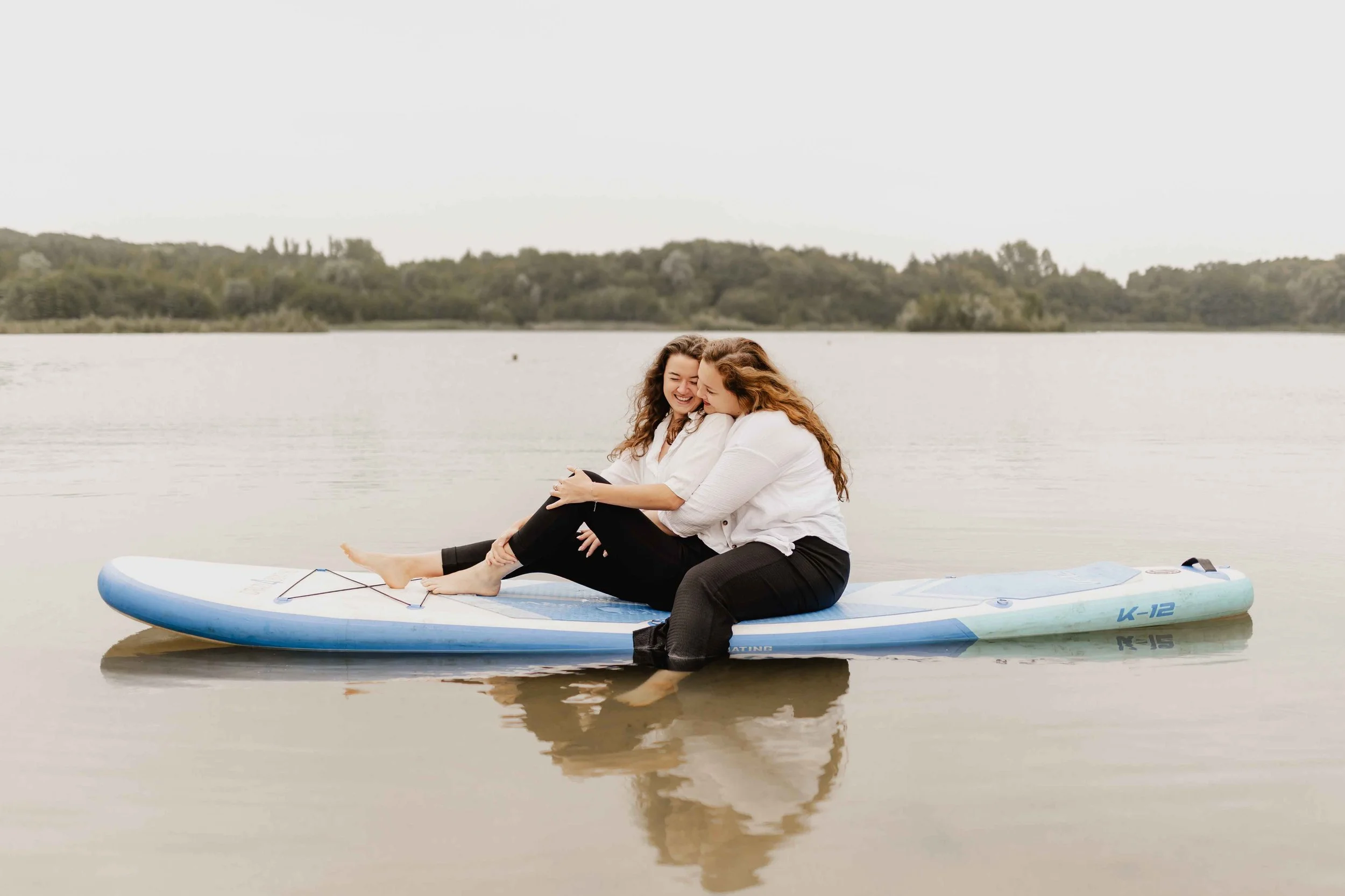 Zwei Frauen sitzen eng umschlungen auf einem SUP-Board auf einem See und lachen