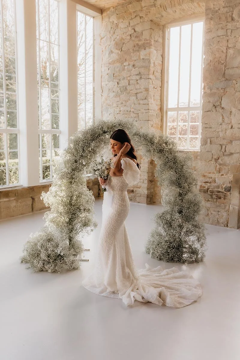 Braut im eleganten Hochzeitskleid vor einem floralen Bogen aus Schleierkraut – moderne und stilvolle Hochzeitsfotografie in Lüneburg mit natürlichen Hochzeitsdetails