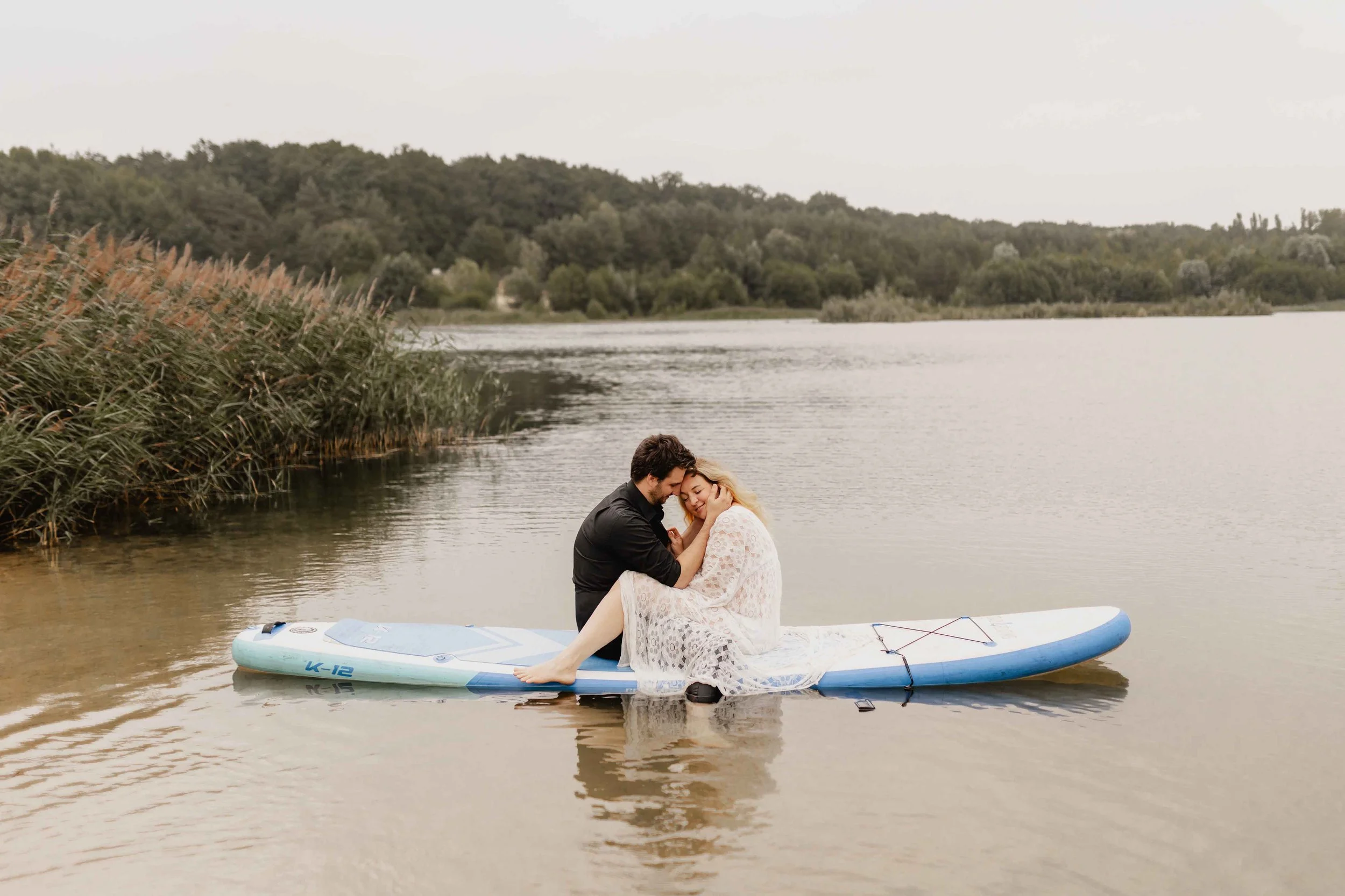 Paar sitzt eng umschlungen auf einem SUP-Board im See mit Bäumen im Hintergrund