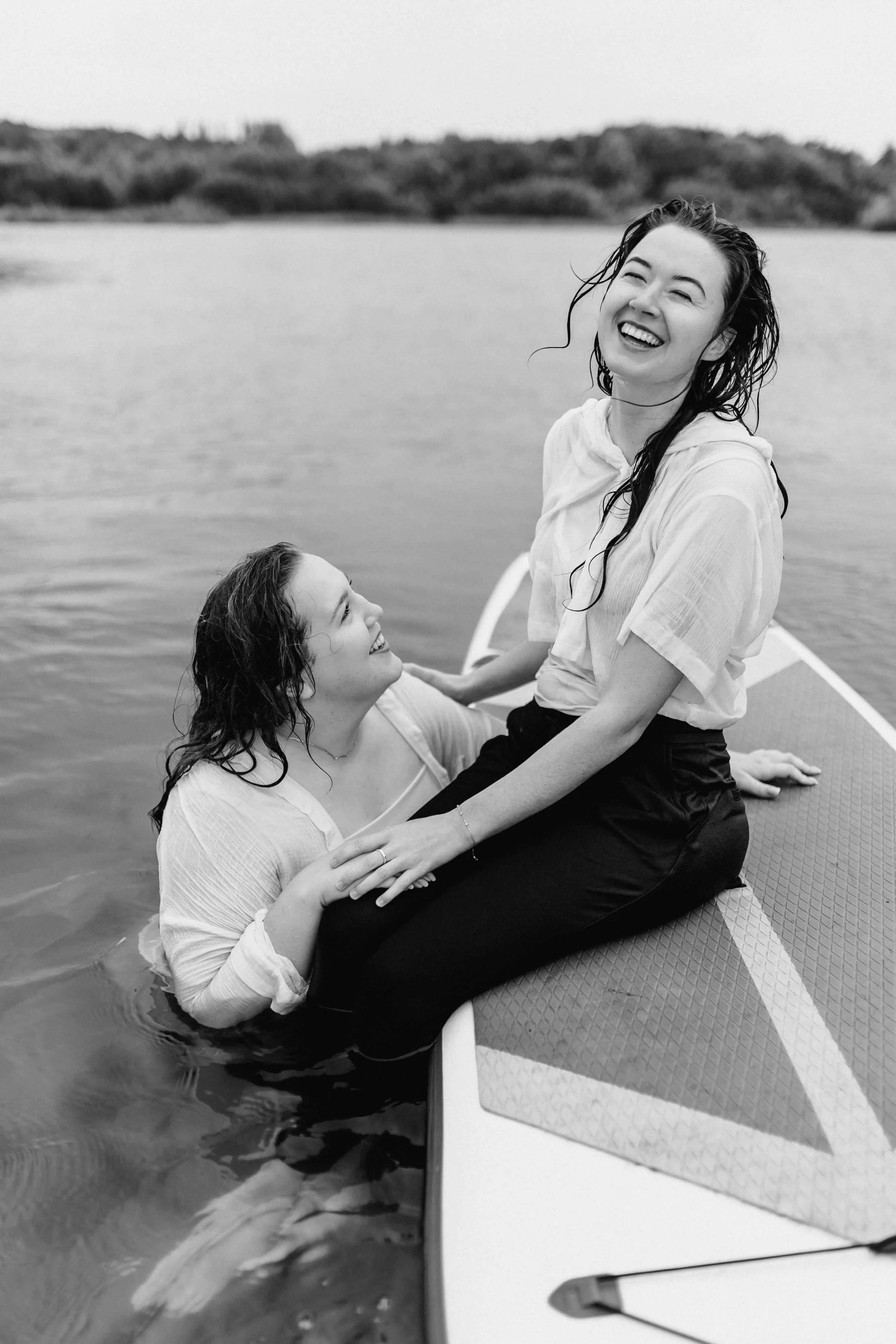 Eine Frau sitzt lachend auf dem SUP, ihre Partnerin hält sie liebevoll aus dem Wasser fest – romantisches Paarshooting auf dem See