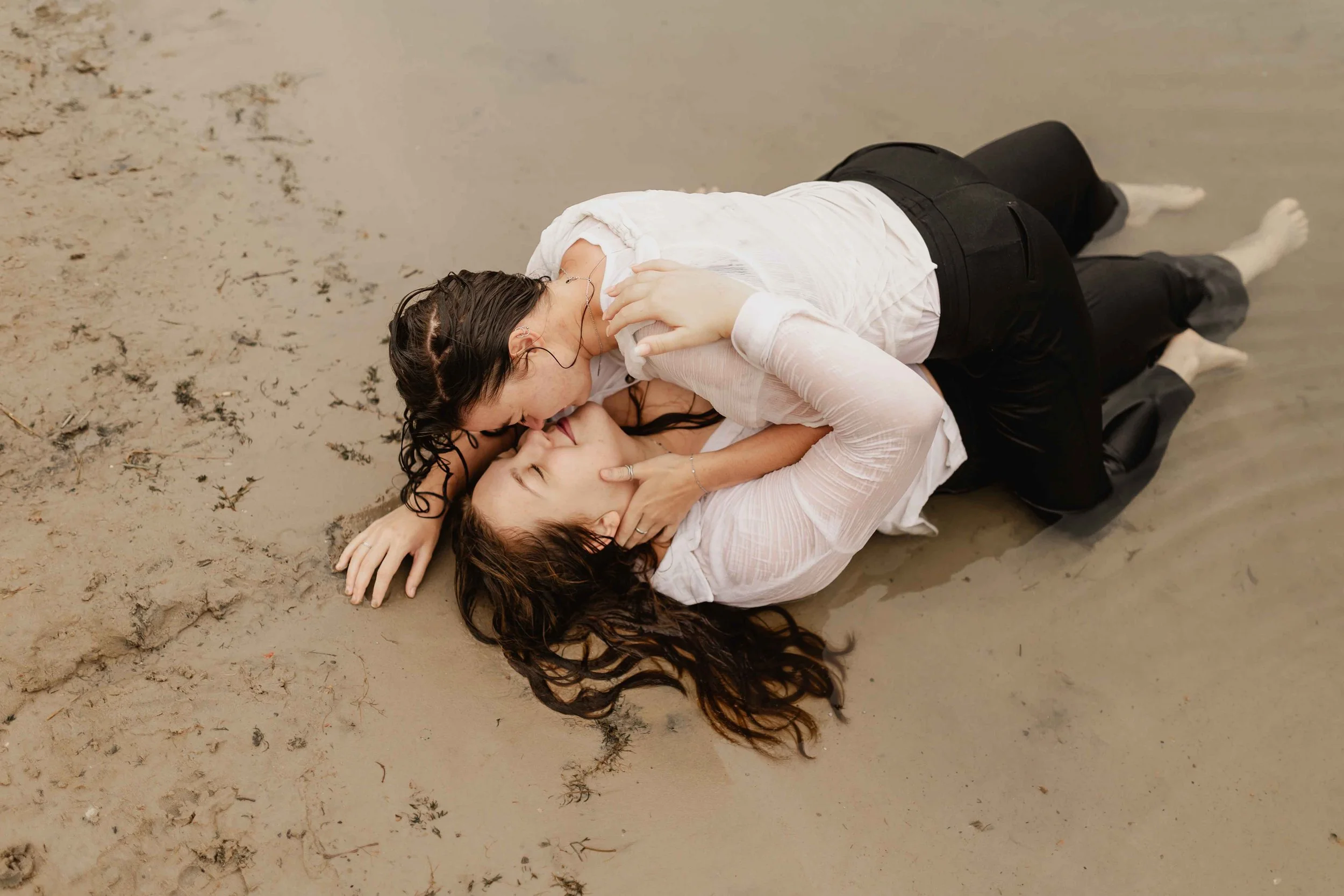 Zwei Frauen liegen eng umschlungen im Sand am See und küssen sich leidenschaftlich