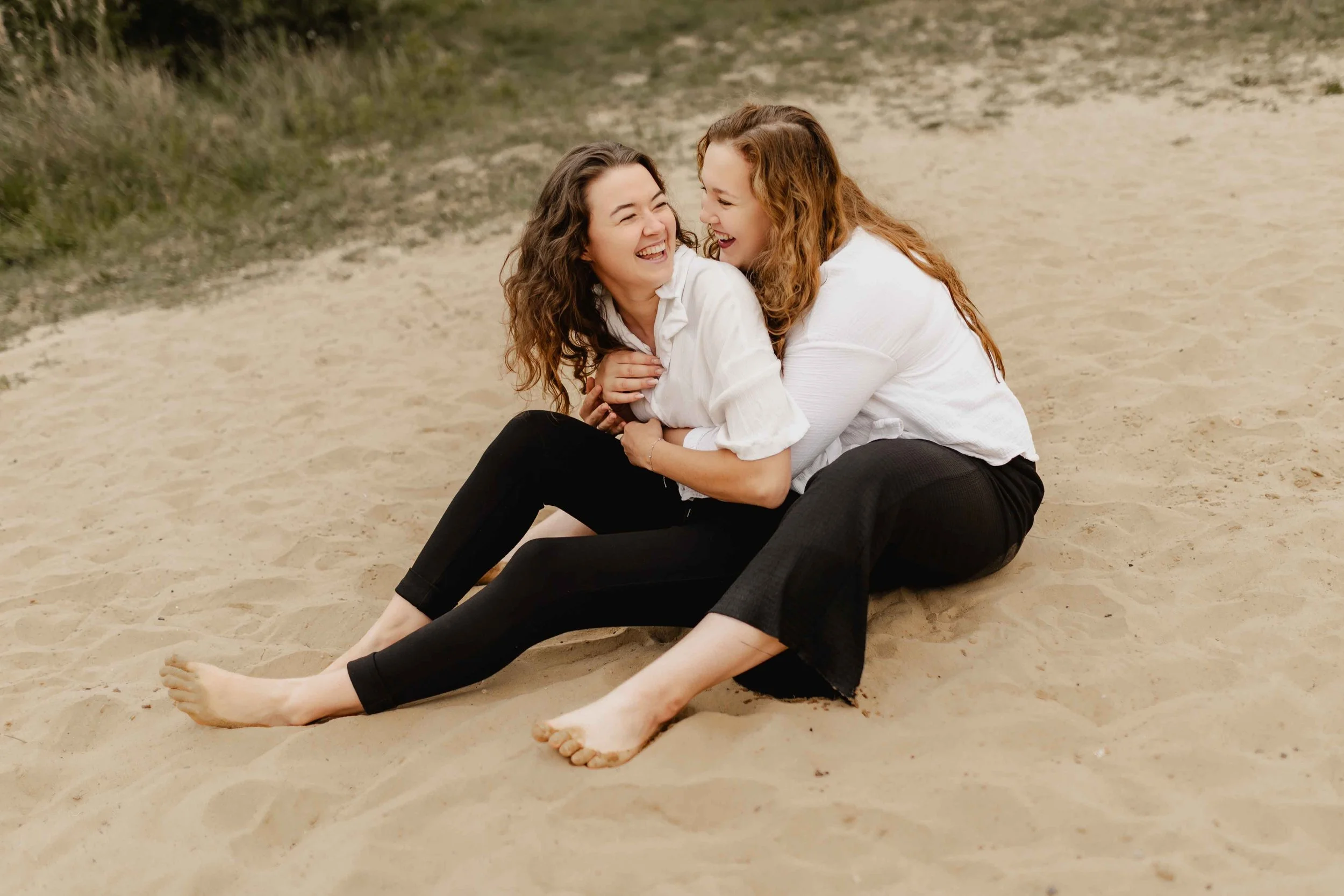 Zwei Frauen sitzen lachend im Sand, eng umschlungen und voller Lebensfreude