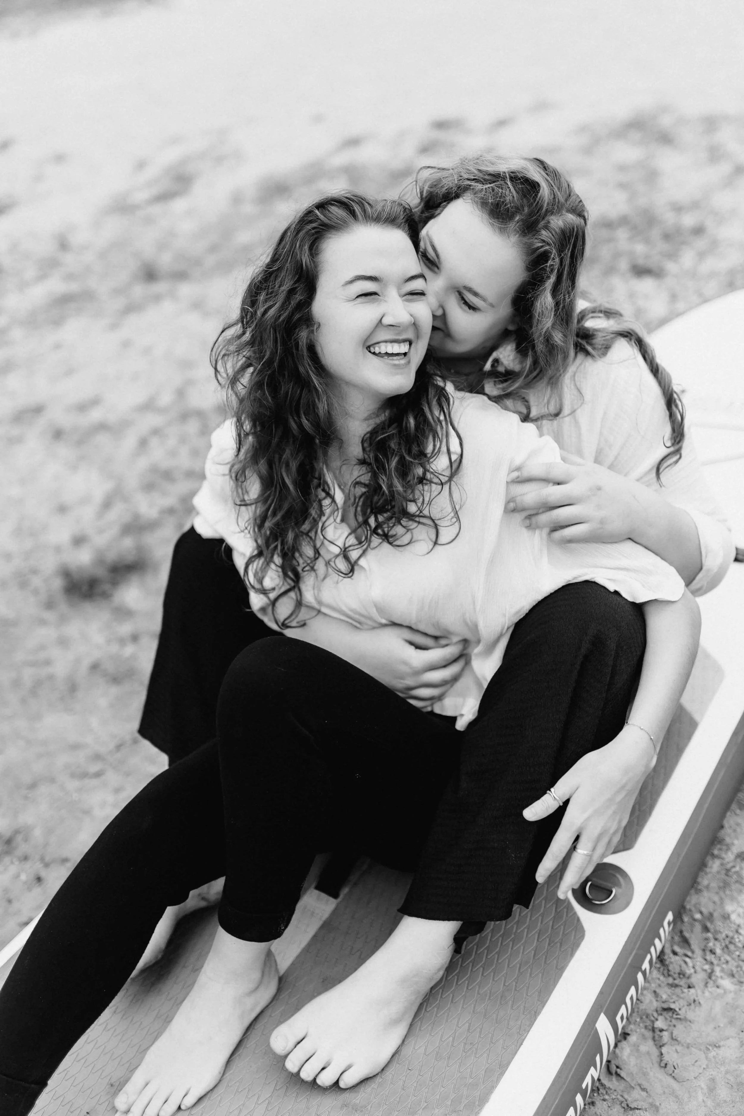 Schwarz-Weiß-Aufnahme von zwei Frauen, die auf einem SUP-Board sitzen, sich umarmen und herzlich lachen