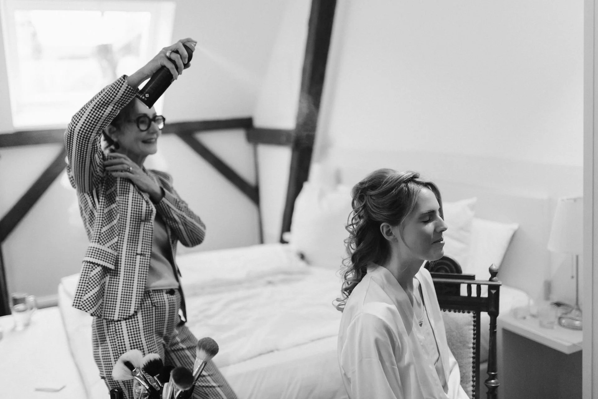 Schwarz-Weiß-Aufnahme einer Braut beim Getting Ready, während die Stylistin ihre Hochzeitsfrisur mit Haarspray fixiert – authentische Hochzeitsreportage im Heide Hotel Reinstorf bei Lüneburg.